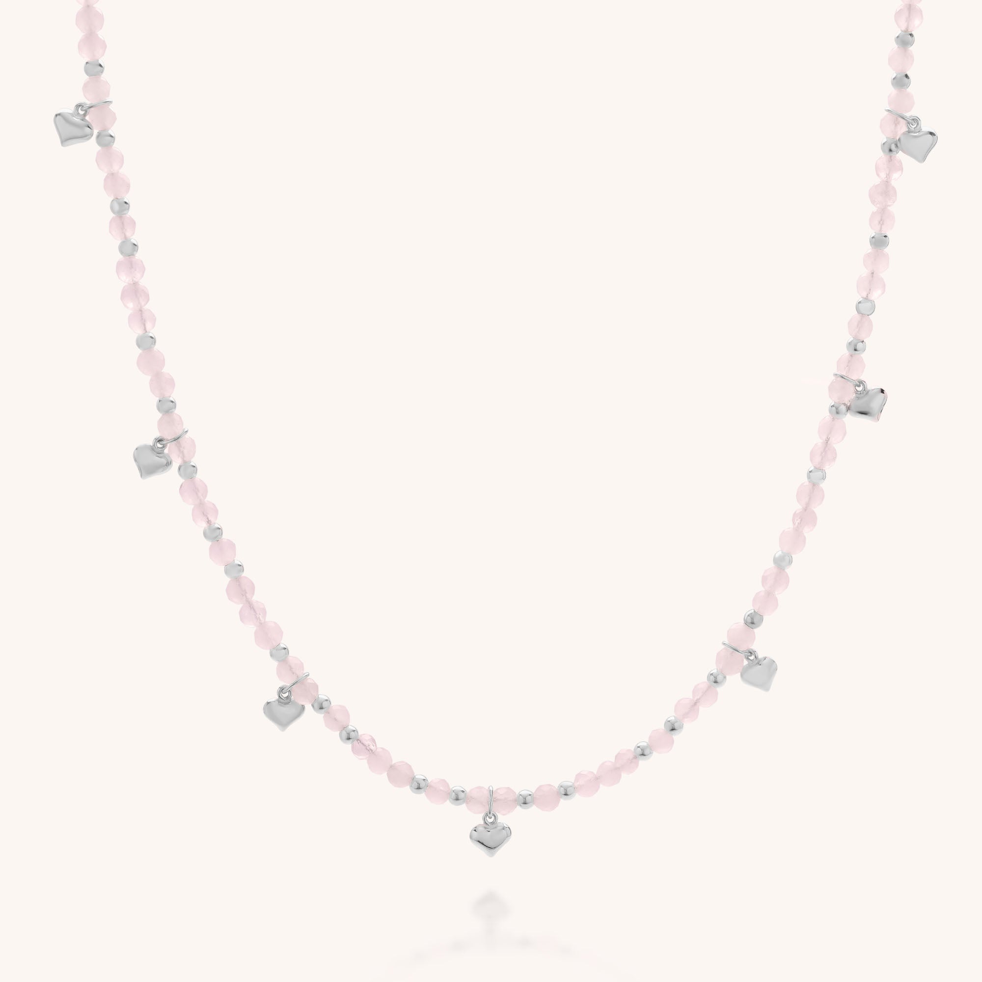 The Rose Hearts Necklace features delicate silver heart charms and small pink beads, beautifully arranged and displayed on a light, neutral background.