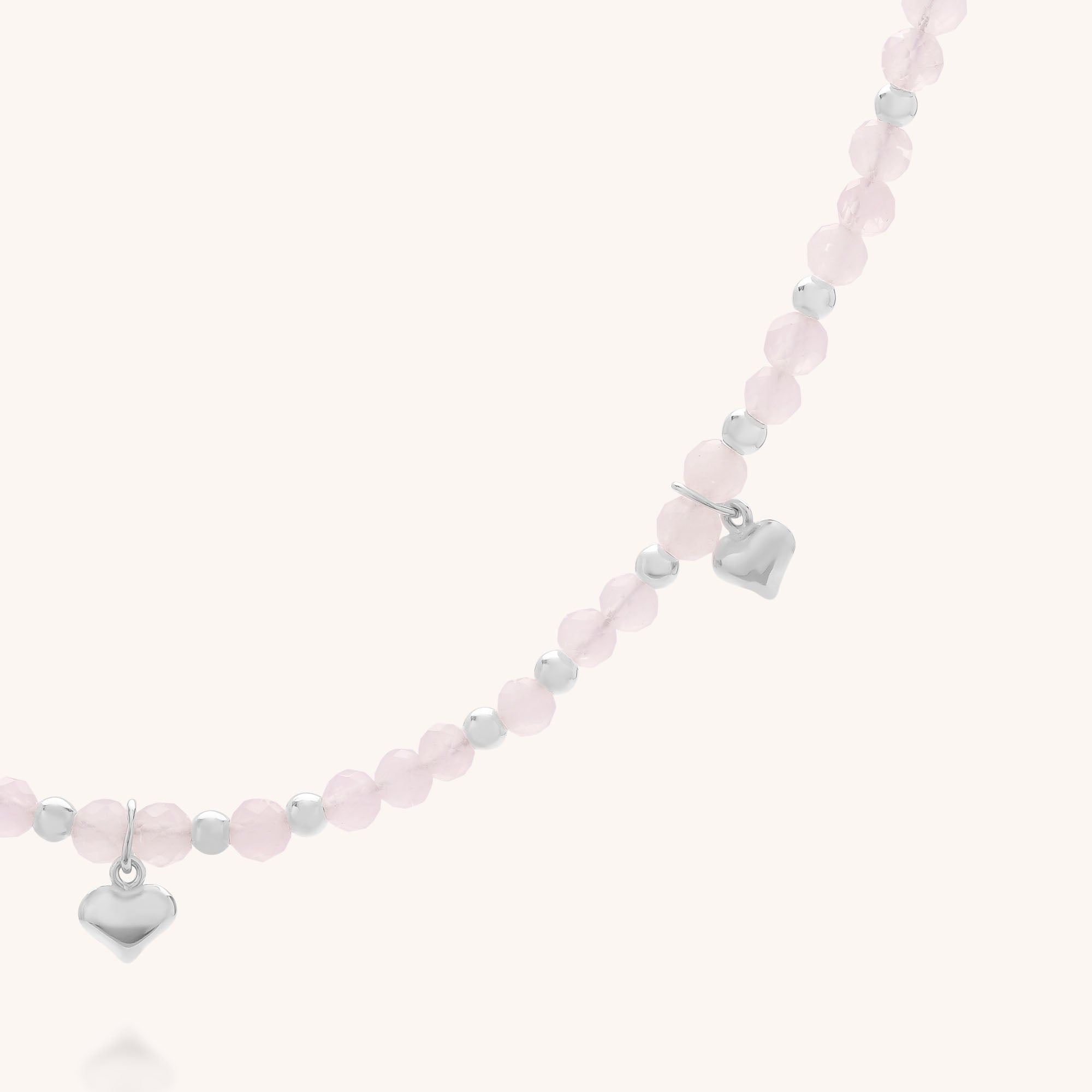 The Rose Hearts Necklace features a delicate strand with alternating light pink and silver round beads, accented by two small silver heart charms that hang from the beaded necklace against a light background.