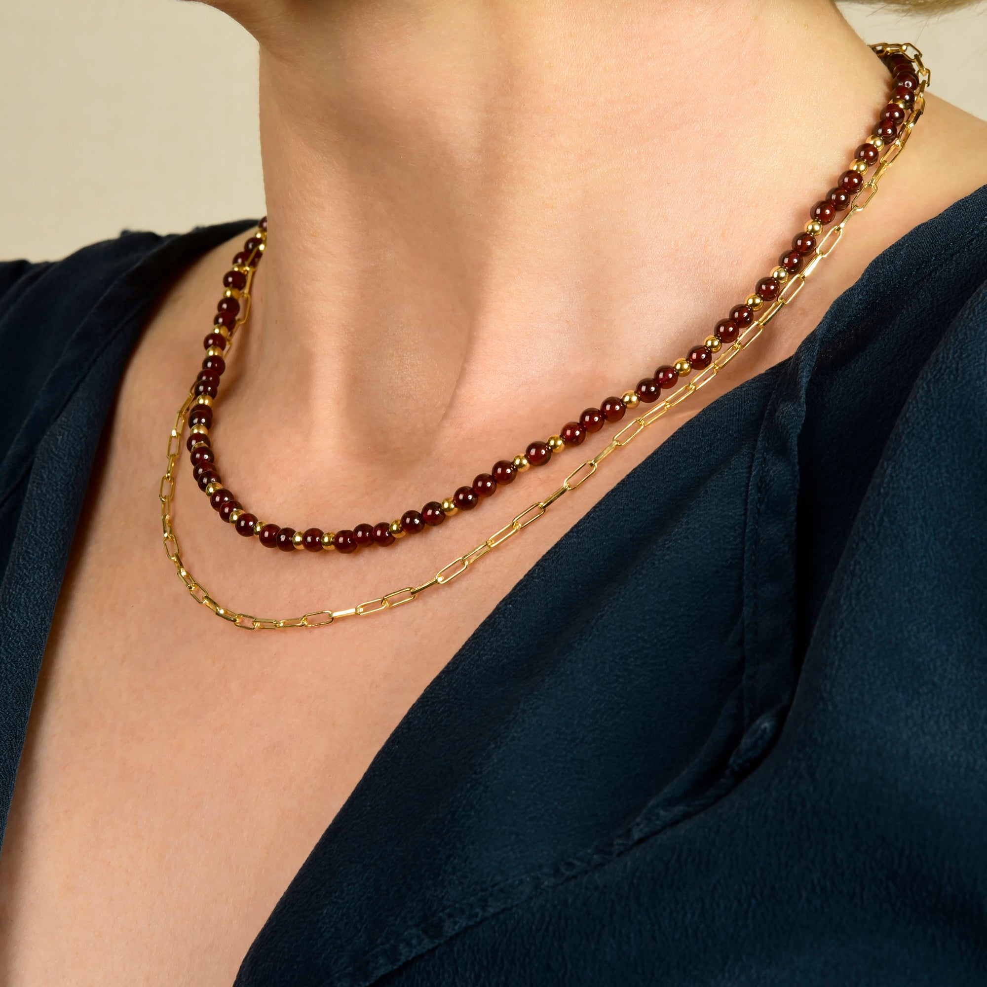A person wearing a navy blue blouse and two necklaces: one featuring eco silver with small red beads and gold accents, the other the Baby Paperclip Chain Necklace in gold.
