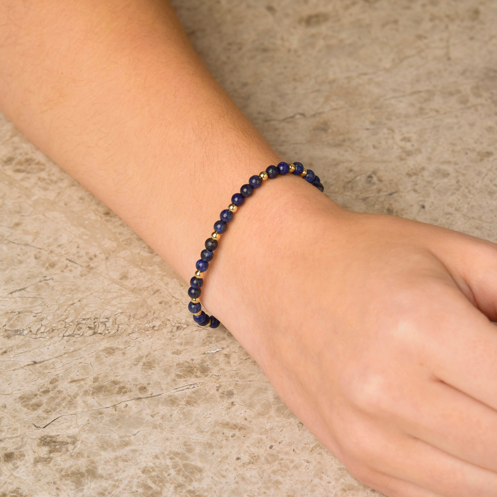 A hand rests on a stone surface, adorned with the Lapis Jewel Bead Birthstone Bracelet - September, featuring small, round blue and gold beads.
