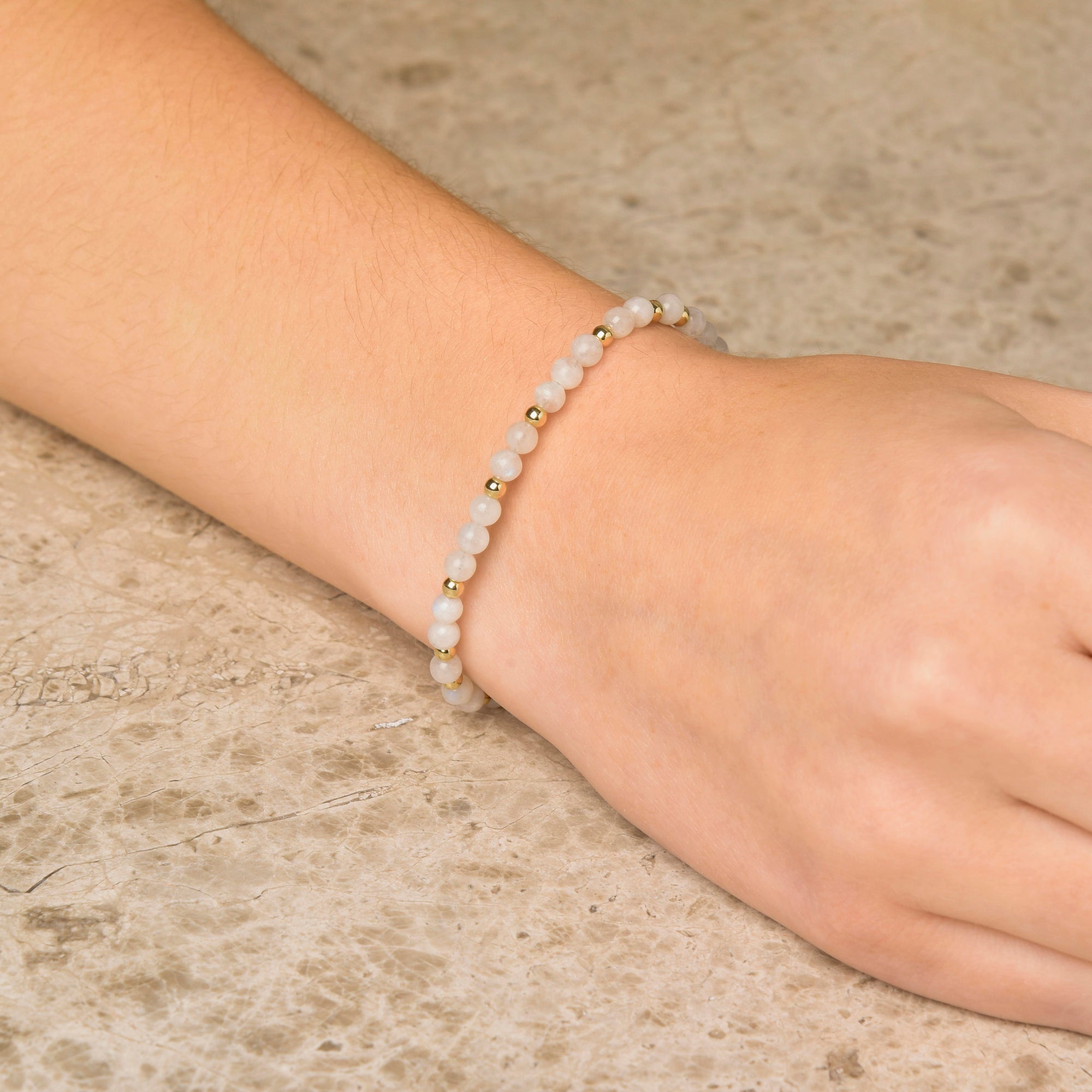 A hand adorned with the Moonstone Jewel Bead Birthstone Bracelet - June, crafted with petite white beads and gold spacers, rests on a textured beige surface—an ideal gift for June birthdays or anyone who loves natural moonstone accents.