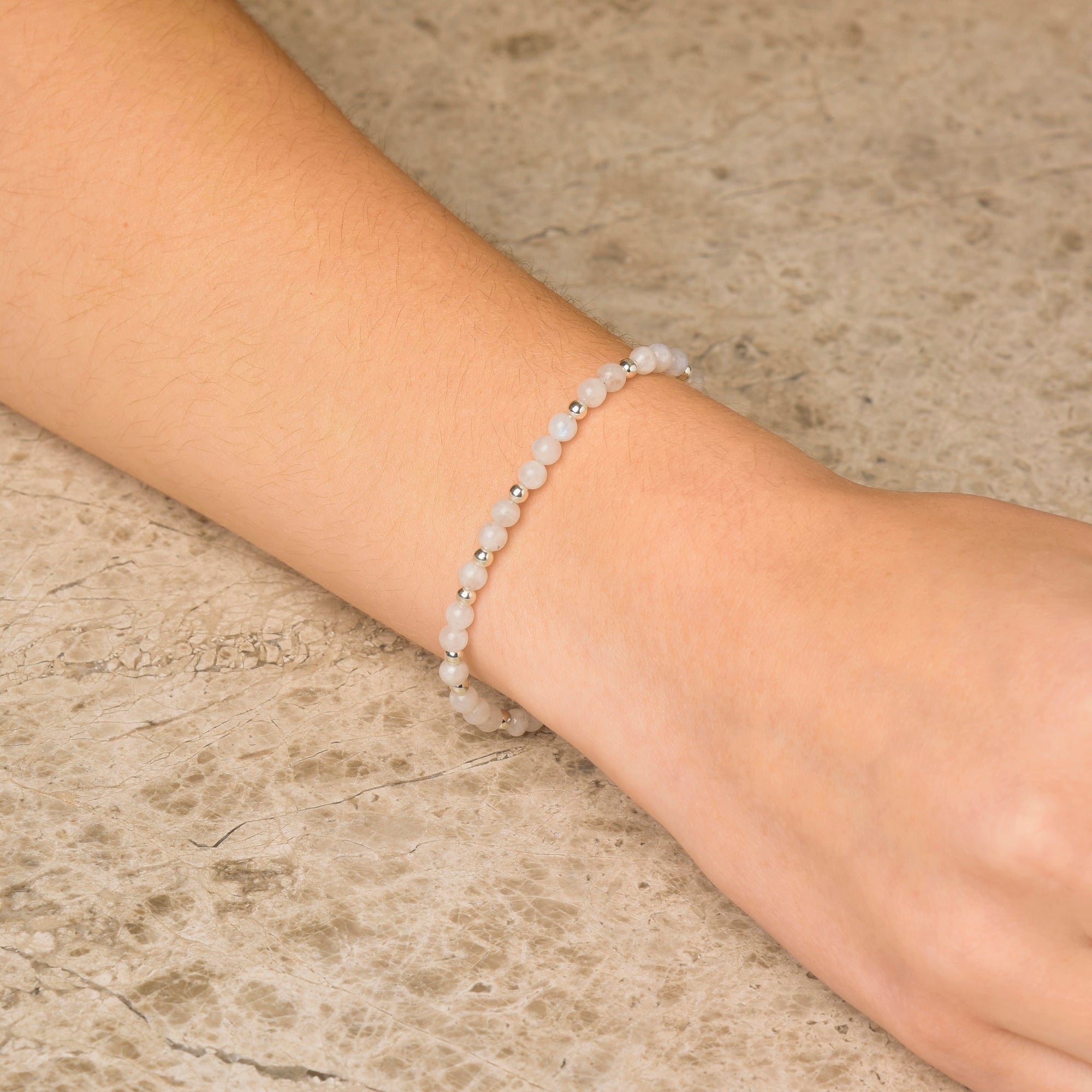 A person wears the Moonstone Jewel Bead Birthstone Bracelet - June on their wrist, showcasing delicate, small round white moonstone beads against a light-colored, textured background. This bracelet is a perfect choice for thoughtful June birthday jewelry.
