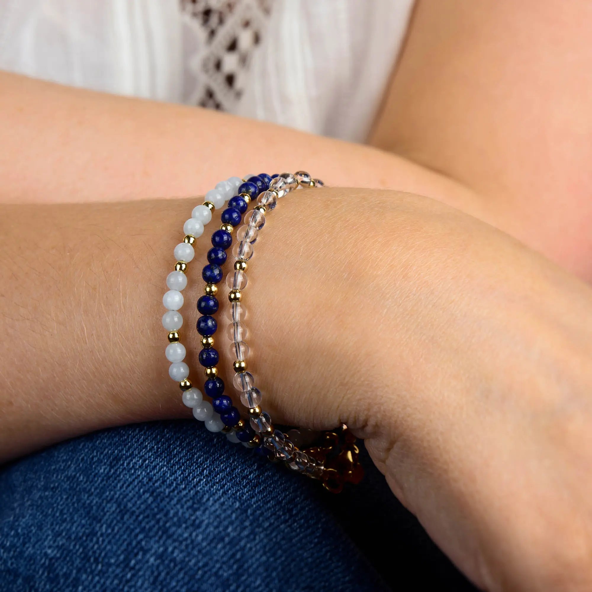 A person’s wrist adorned with four beaded bracelets—including the striking Jewel Bead Birthstone Bracelet featuring white, blue, gold, and clear beads—rests on their lap while a white lace top peeks through. Handcrafted in the UK, this unique accessory adds a charming touch to any outfit and is perfect for those who appreciate distinctive jewelry pieces.