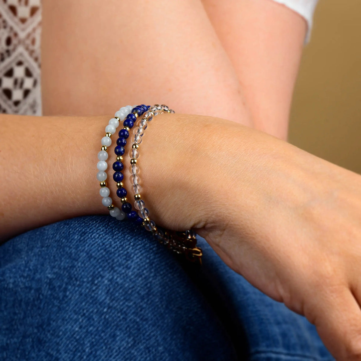 A person wearing three beaded bracelets—including the eye-catching Lapis Jewel Bead Birthstone Bracelet - September—in blue, white, and gold tones on their wrist, rests their arm on their knee. They are dressed in blue jeans and a white patterned top.