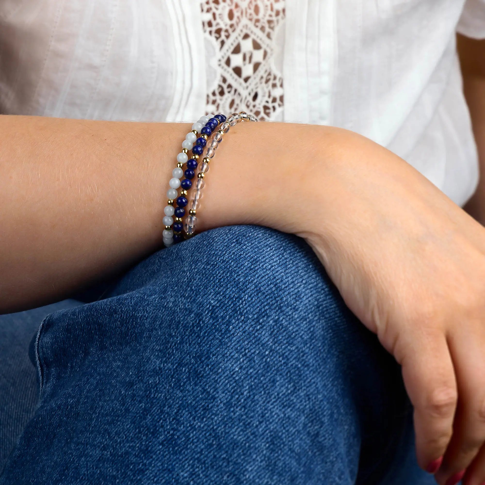 A person in a white shirt and blue jeans rests their left arm on their knee, adorned with three layered beaded bracelets in blue, white, and gold tones, including the Moonstone Jewel Bead Birthstone Bracelet - June, which features delicate natural moonstone accents.
