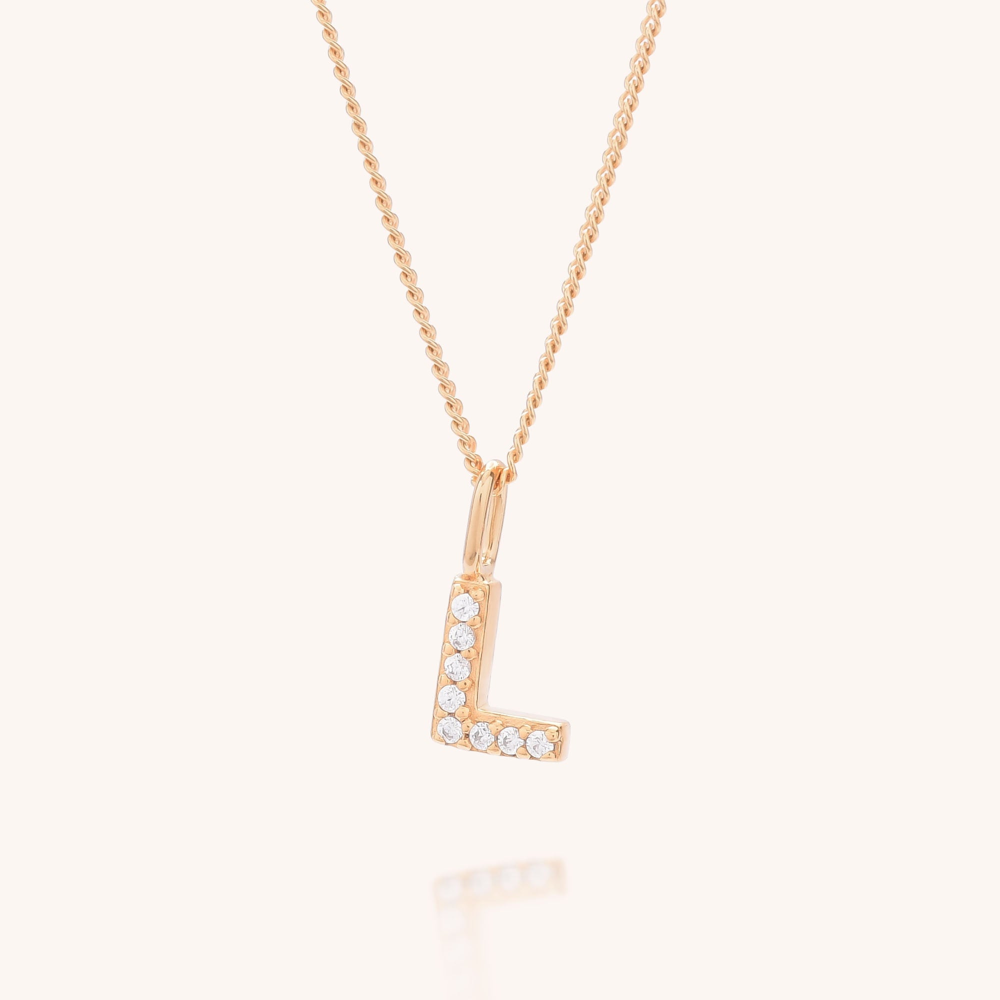 The Initial Sparkle Necklace is a gold necklace featuring an L-shaped pendant adorned with sparkling cubic zirconia gems, shown against a light cream background.