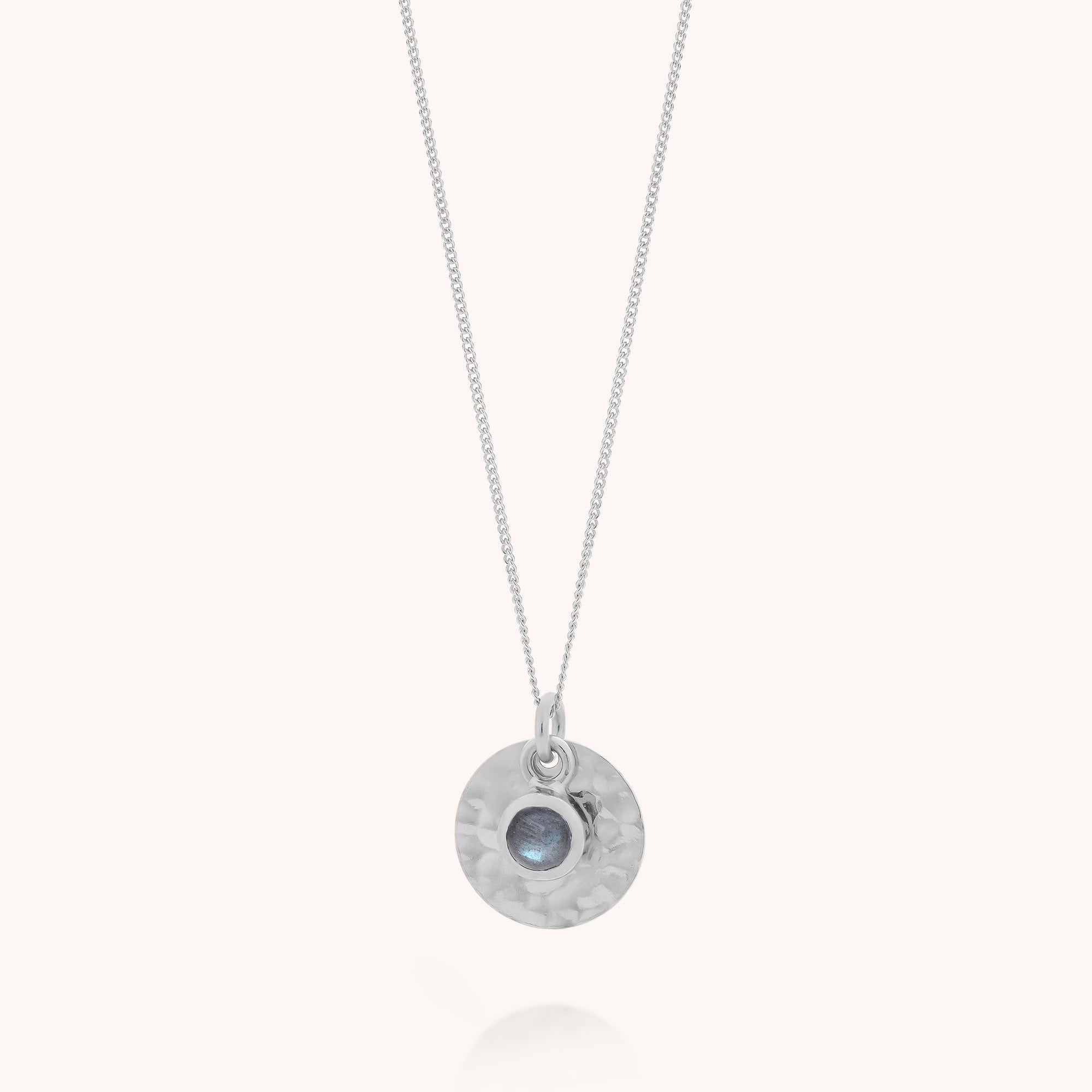The Labradorite Luna Disc Necklace is crafted from sterling silver and features a fine chain with a round, hammered pendant set with a small, blue-gray labradorite stone at its center, displayed against a simple light background.