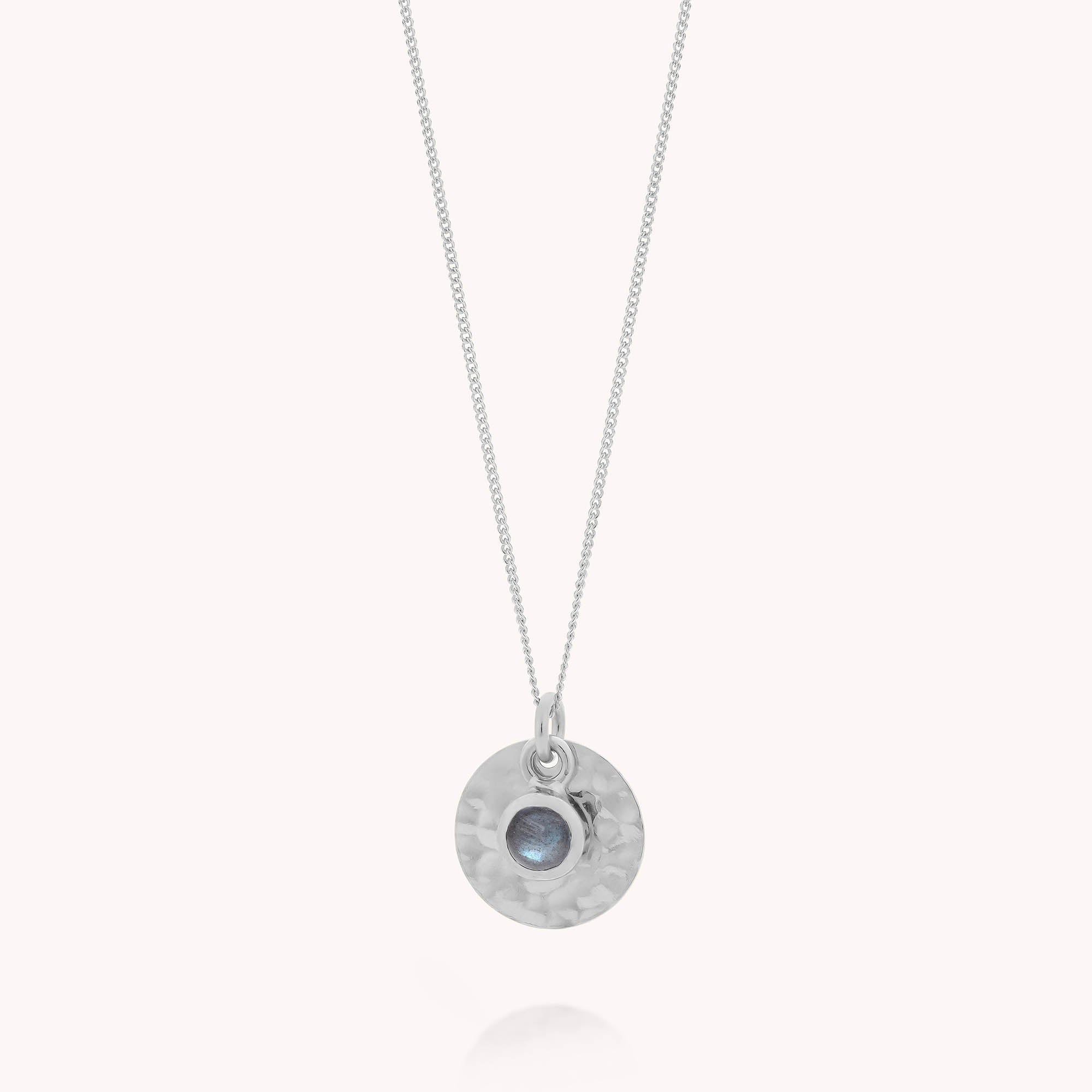 The Labradorite Luna Disc Necklace is a sterling silver pendant with a hammered round disc, featuring a central blue labradorite stone, suspended from a fine silver chain.