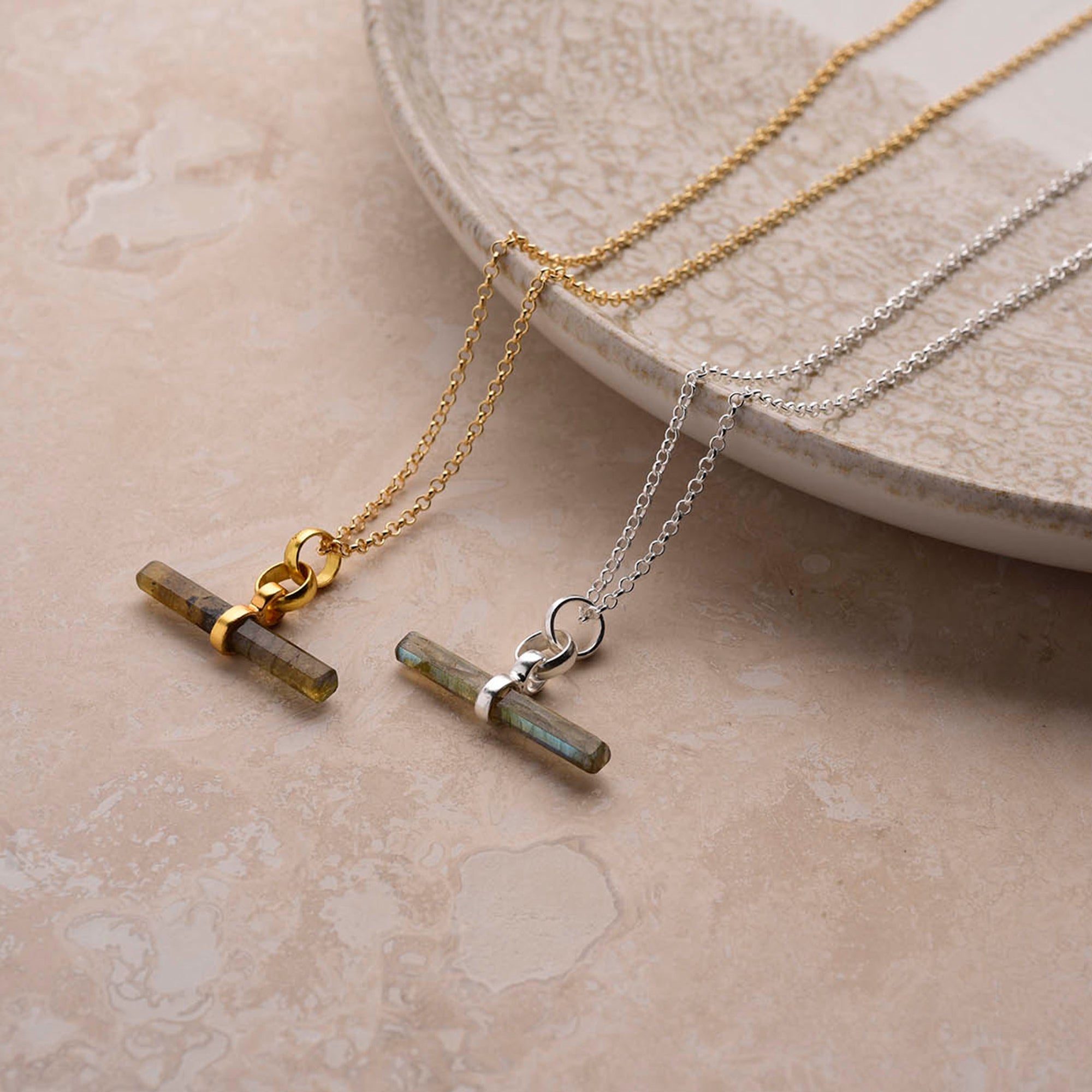 Two Gemstone T Bar Necklaces—one with a gold chain, one with a silver chain—are displayed on a beige stone surface beside a ceramic plate.