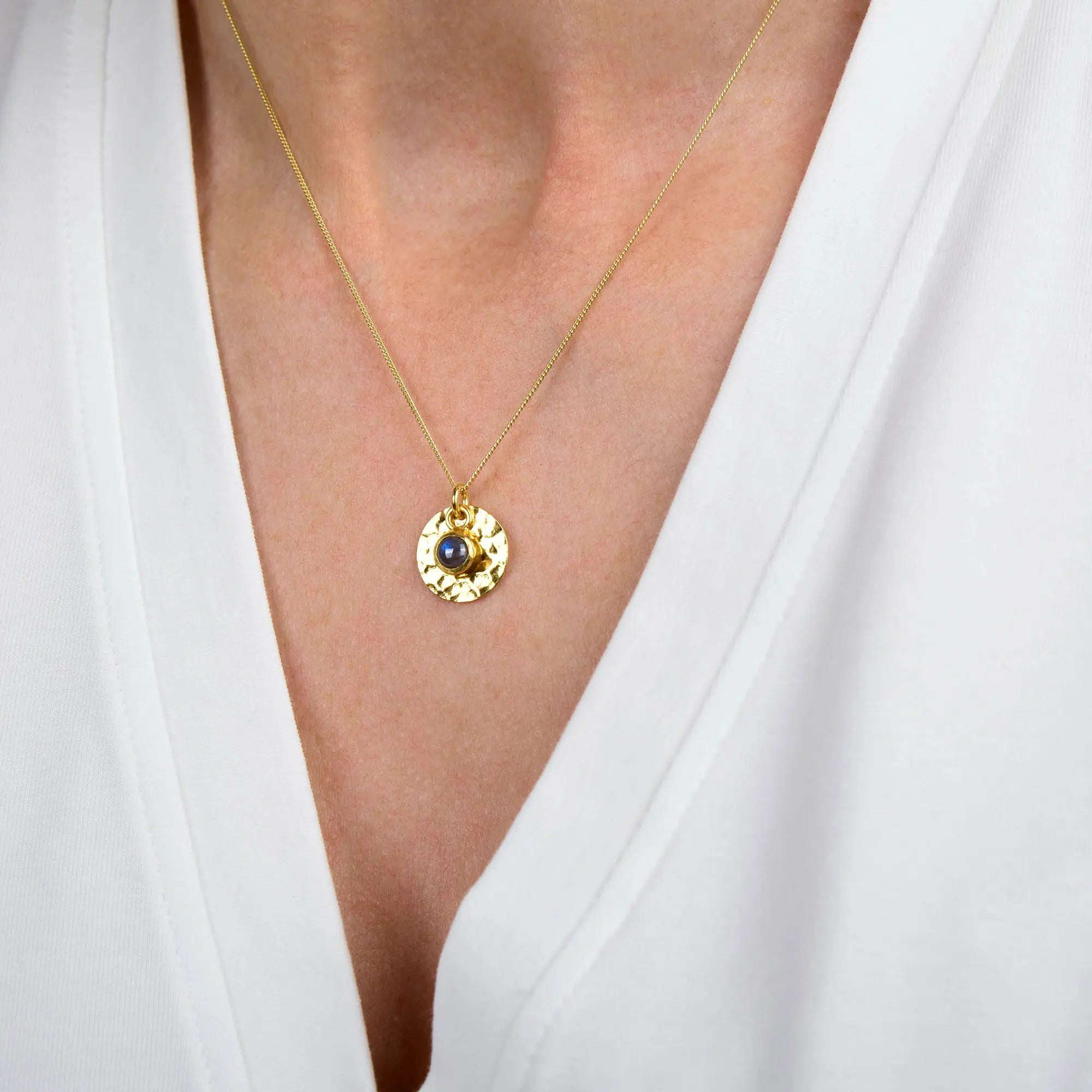 The Labradorite Luna Disc Necklace, featuring a textured gold round pendant and a striking blue labradorite gemstone, is worn around the neck and stands out against a white V-neck shirt.