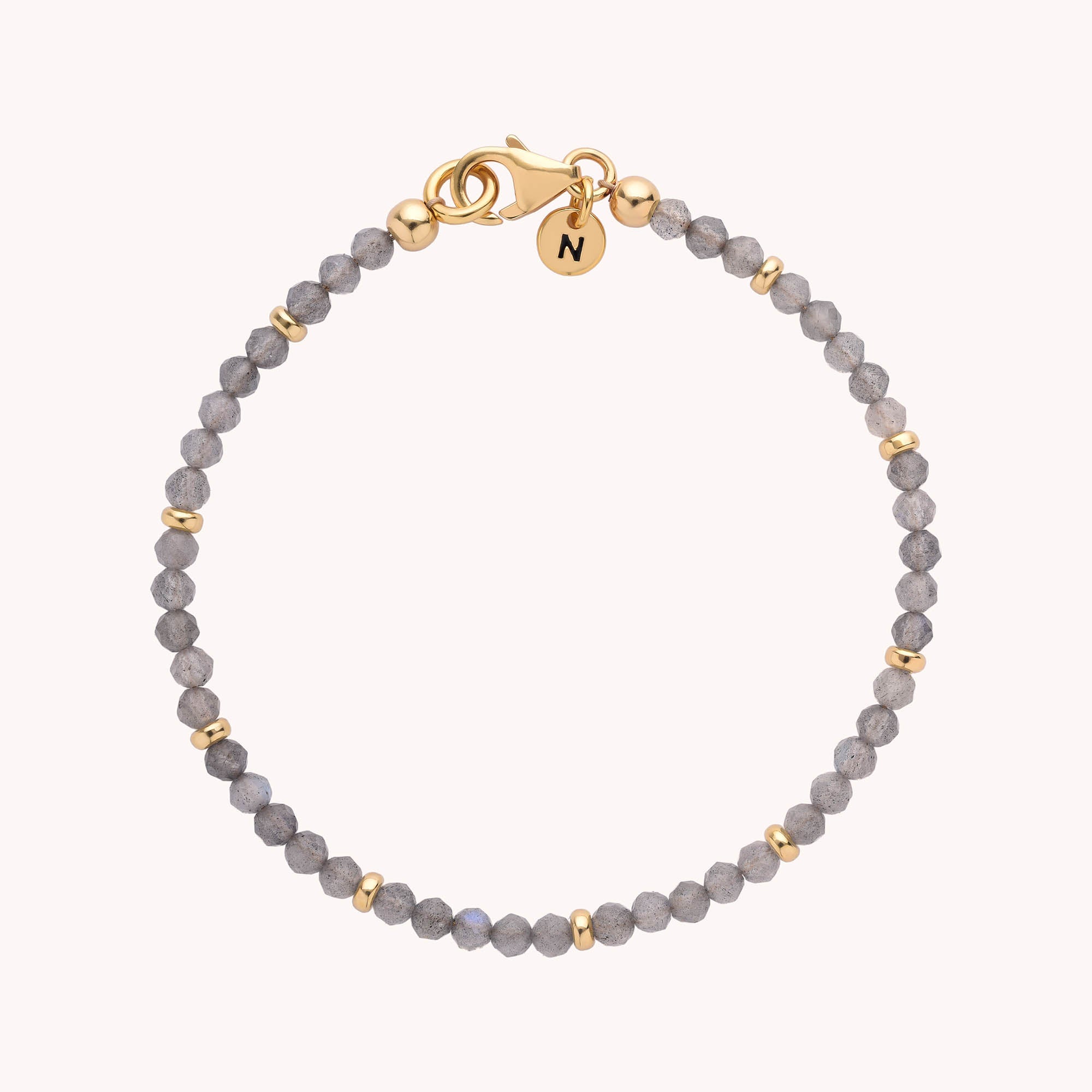 Presenting the Labradorite Orbit Bracelet: a beaded piece adorned with small gray labradorite stones and elegant gold accents, featuring a gold clasp and a round gold charm engraved with the letter "N.
