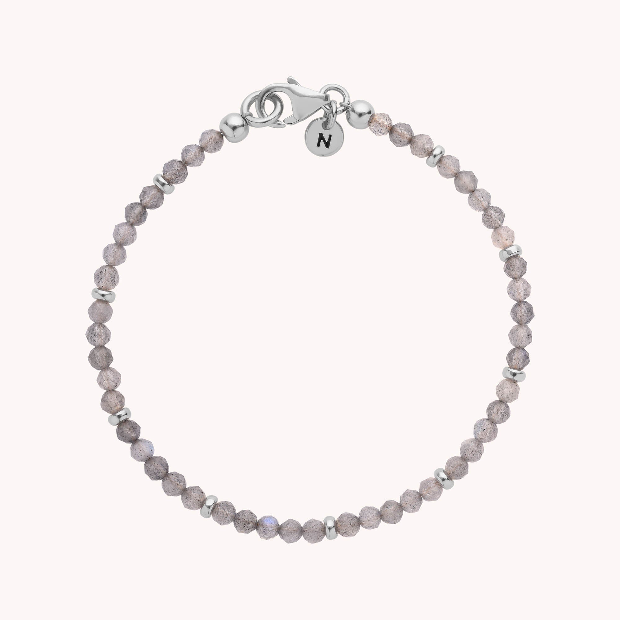 The Labradorite Orbit Bracelet is a delicate sterling silver piece adorned with light purple labradorite stones, small silver beads, a secure silver clasp, and finished with a round charm engraved with the letter N, beautifully displayed on a plain light background.