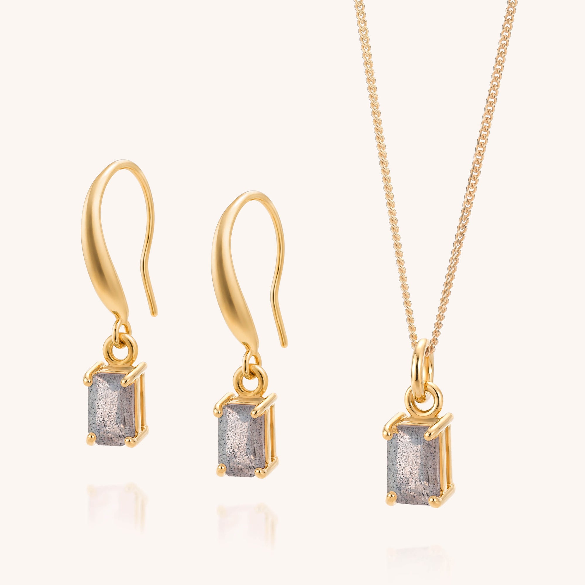 The Labradorite Octavia Necklace & Earrings Set features gold earrings and a matching gold necklace, each adorned with a rectangular gray labradorite gemstone in a classic prong setting. Displayed on a light background, this elegant set combines timeless sophistication with the captivating charm of labradorite.