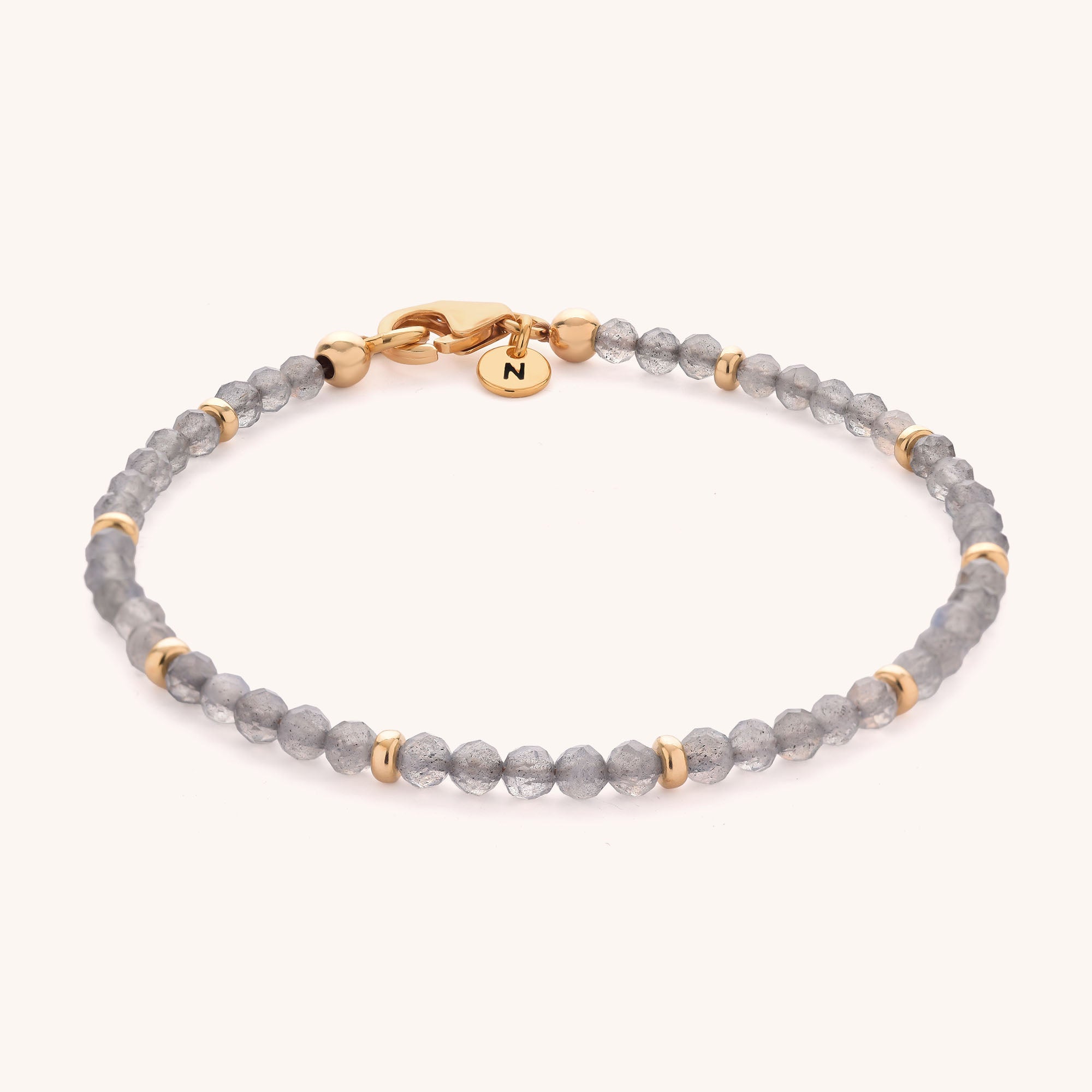 The Labradorite Orbit Bracelet features faceted gray labradorite beads, small gold spacer beads, a gold clasp, and a round gold charm engraved with the letter N.