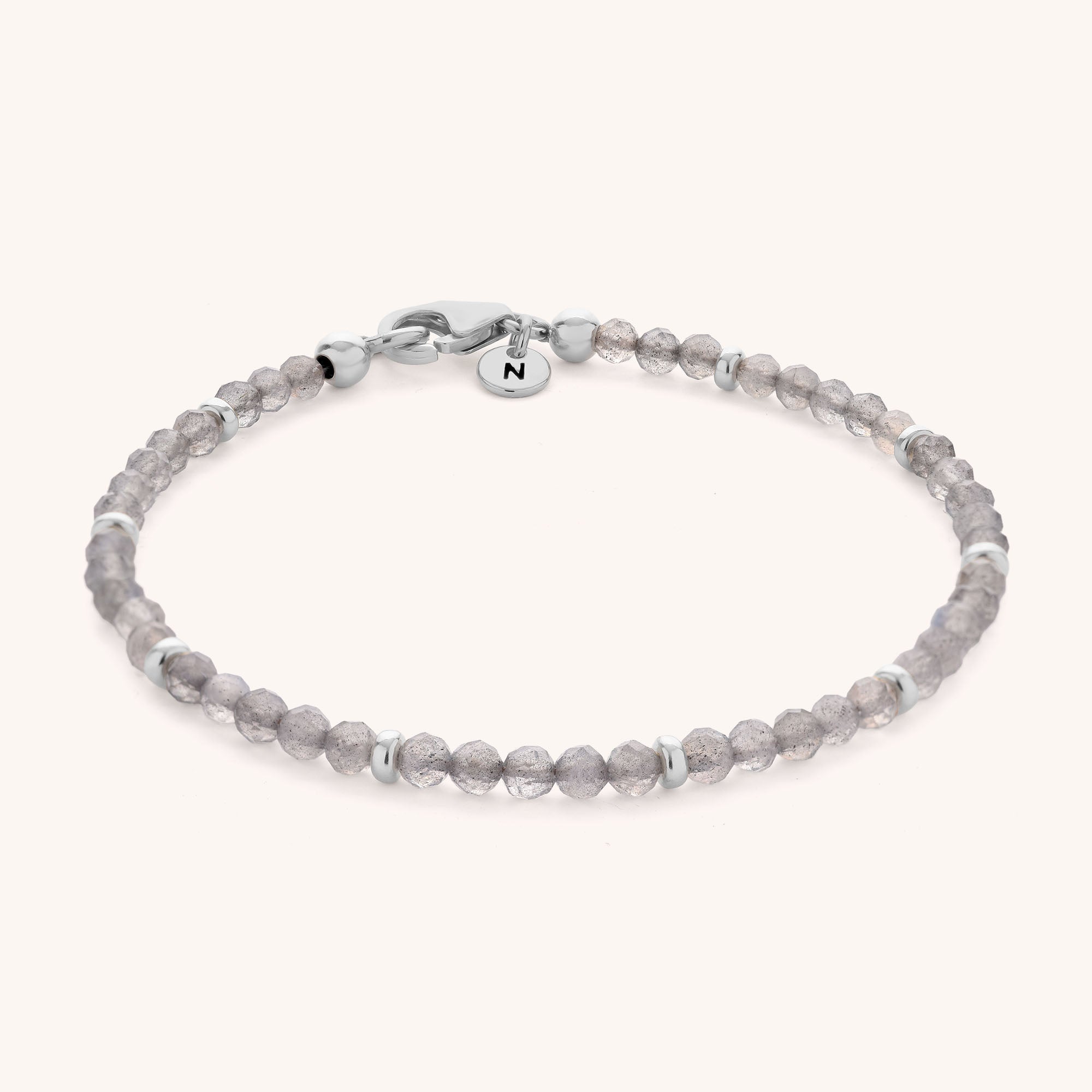 Labradorite Orbit Bracelet featuring small, round, faceted grey labradorite beads paired with silver accent beads, finished with a sterling silver clasp and a small circular charm engraved with the letter N on a light background.