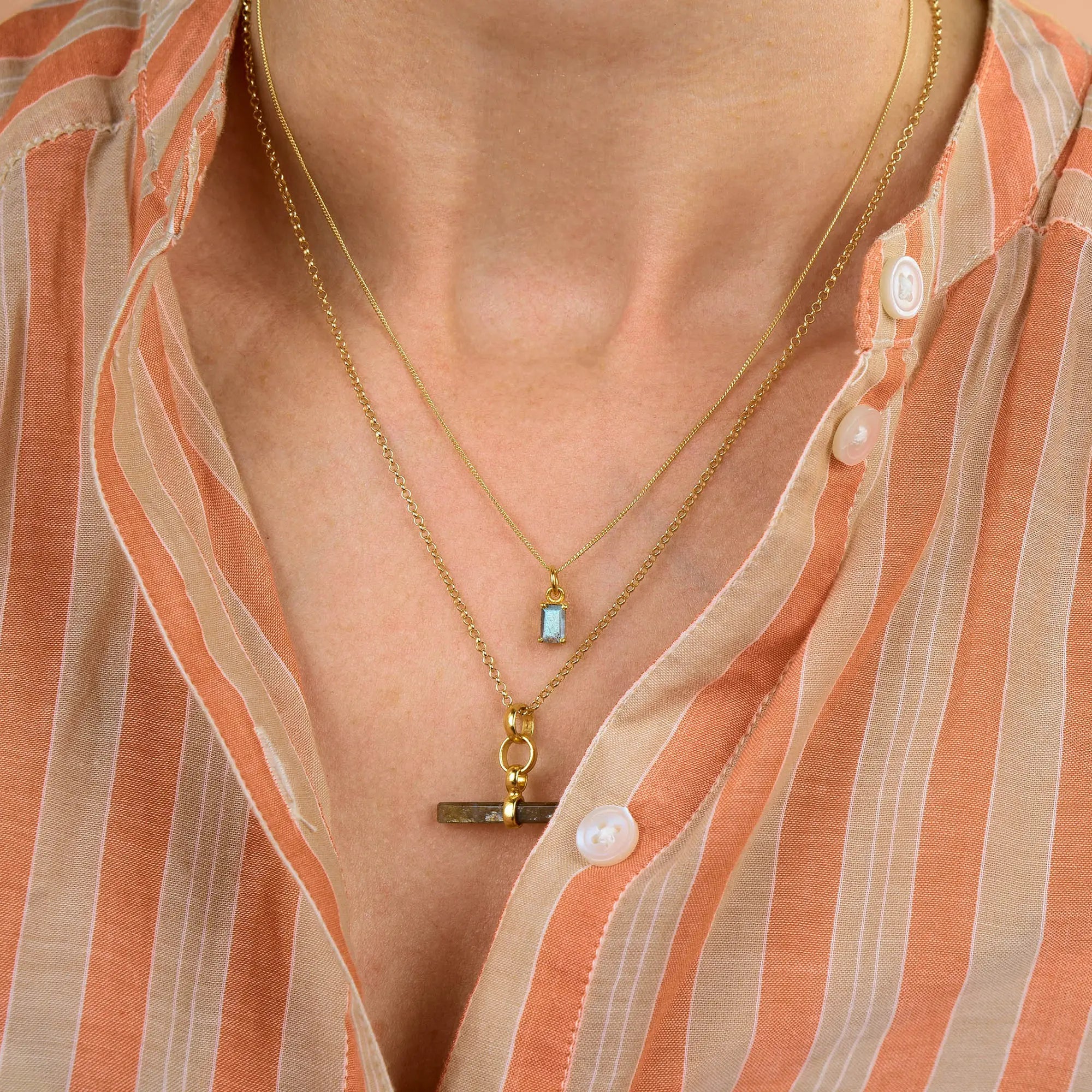 A person wearing a peach and beige striped shirt with two gold necklaces: one featuring the Labradorite Octavia Necklace with a small rectangular labradorite pendant, and the other adorned with a horizontal gold bar pendant.