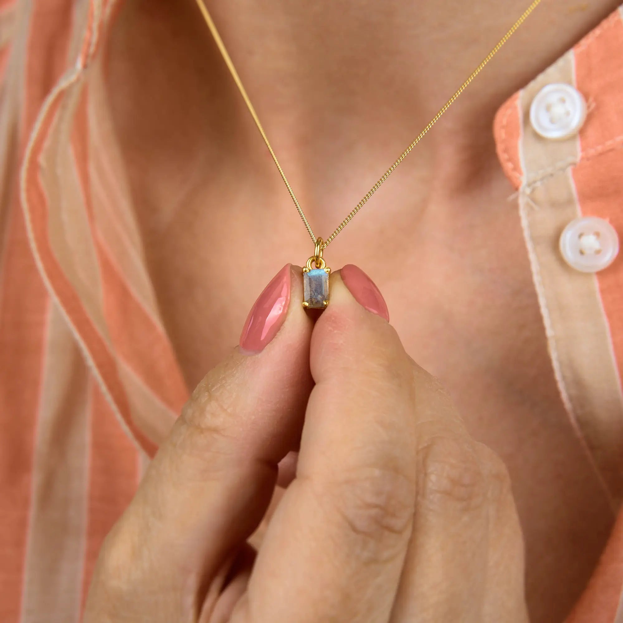 A person with pink nail polish holds the Labradorite Octavia Necklace, a gold octagonal design featuring a rectangular labradorite gemstone pendant, and wears a peach and white striped shirt with white buttons.