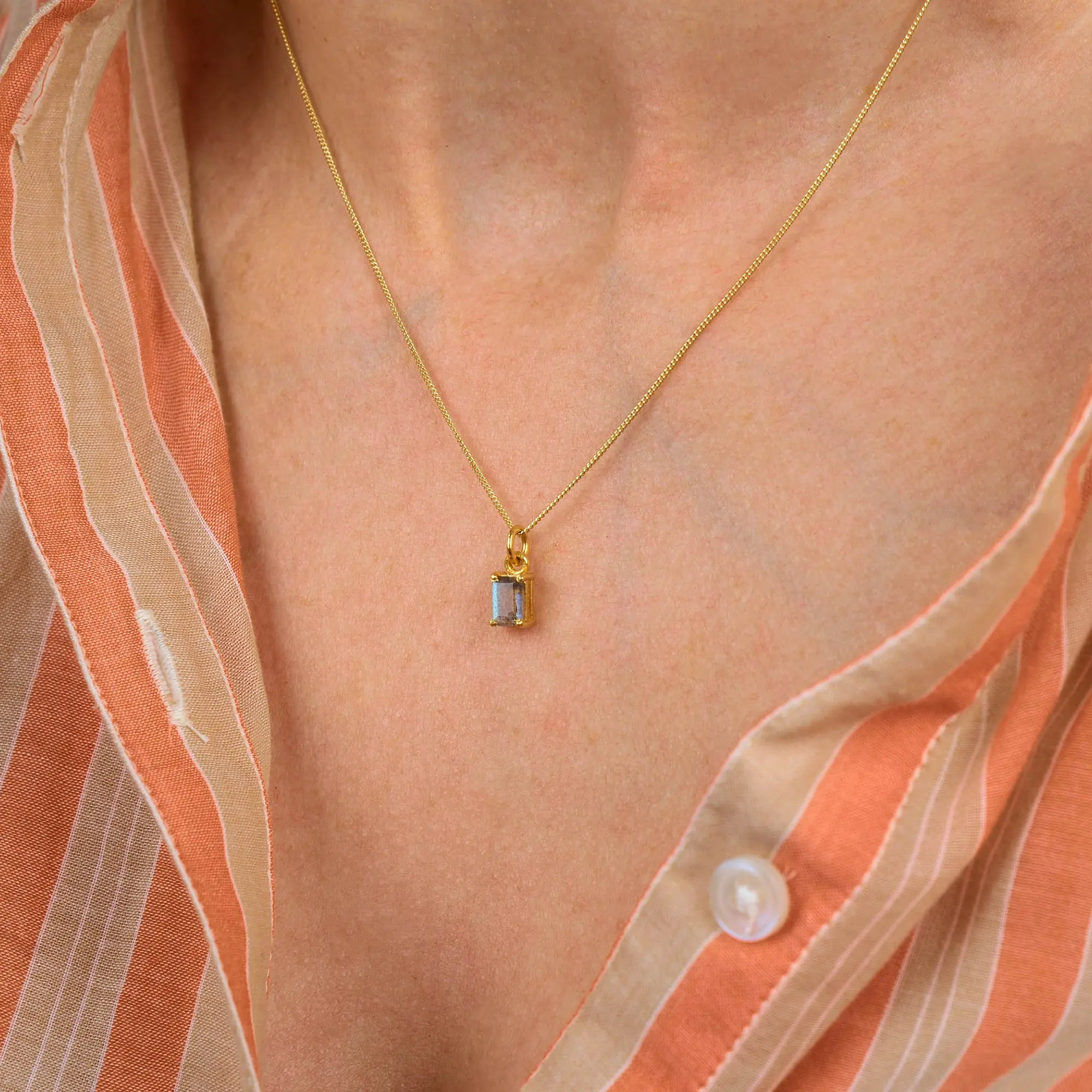 A close-up of a person wearing the Labradorite Octavia Necklace—a gold chain featuring a rectangular labradorite gemstone pendant—paired with a peach and beige striped shirt. The shimmering necklace catches the light beautifully, adding an elegant touch to the ensemble.
