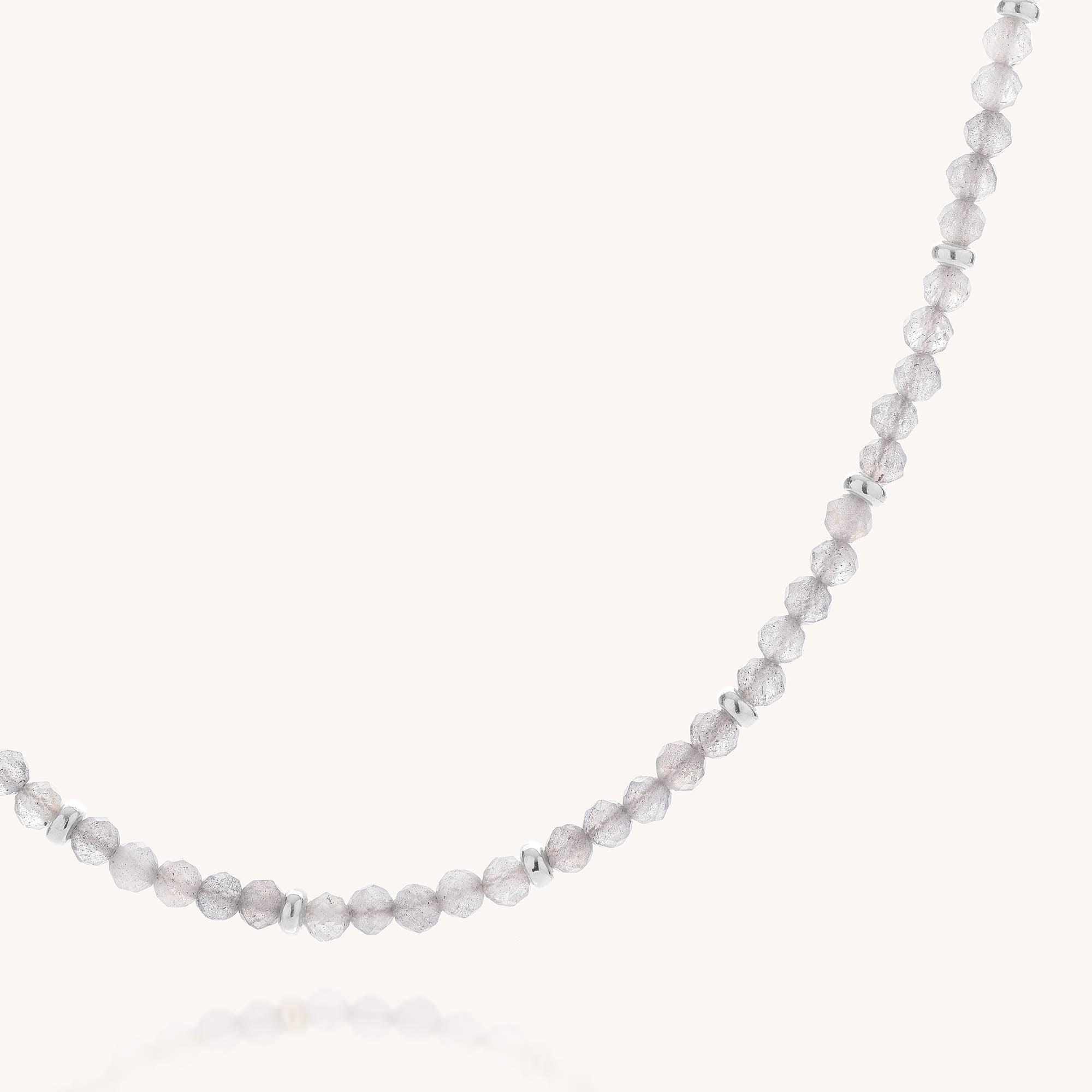 The Labradorite Orbit Necklace features small, round, translucent faceted labradorite beads accented with shiny sterling silver spacers, artfully arranged in a gentle curve against a plain light background.