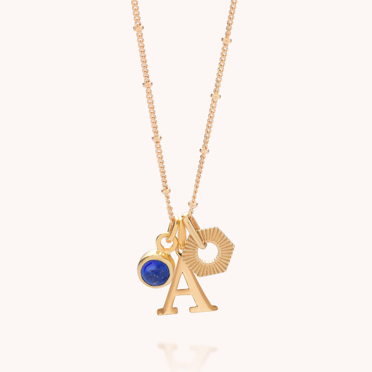 Introducing the HEX Personalised Birthstone Necklace – Gold Plated: a stunning gold-plated sterling silver necklace featuring three unique charms—a blue birthstone gemstone set in a gold bezel, a gold letter A, and a textured gold hexagonal pendant—all suspended from a delicate gold chain. Presented against a light background, this personalised birthstone necklace is the perfect thoughtful gift for any special occasion.
