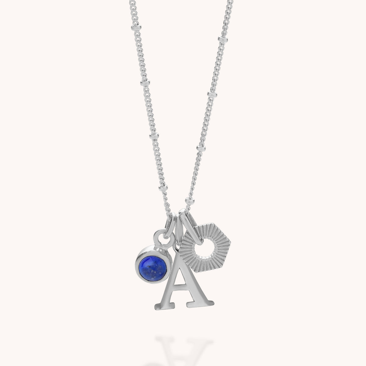 Introducing the HEX Personalised Birthstone Necklace – Sterling Silver: a beautifully crafted necklace showcasing three elegant pendants—a sparkling blue birthstone-inspired circular gem, a customisable capital letter A, and a textured silver hexagon.