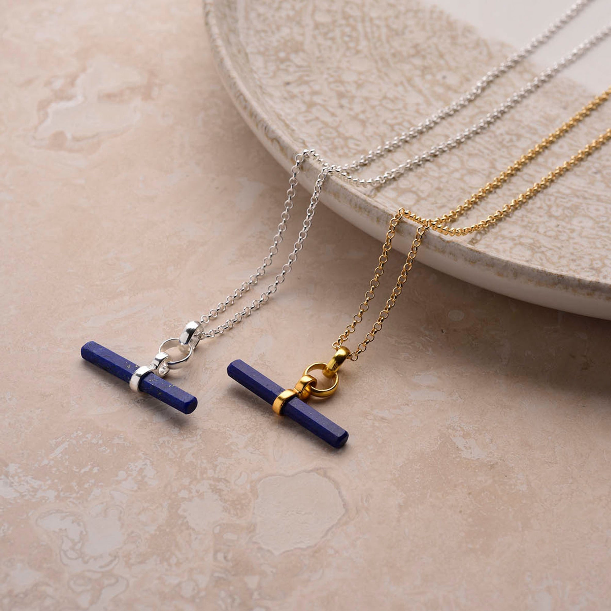 Two Gemstone T Bar Necklaces with blue gemstone pendants rest on beige marble. One has a sterling silver chain, the other features gold. Both pendants display elegant metallic detailing.