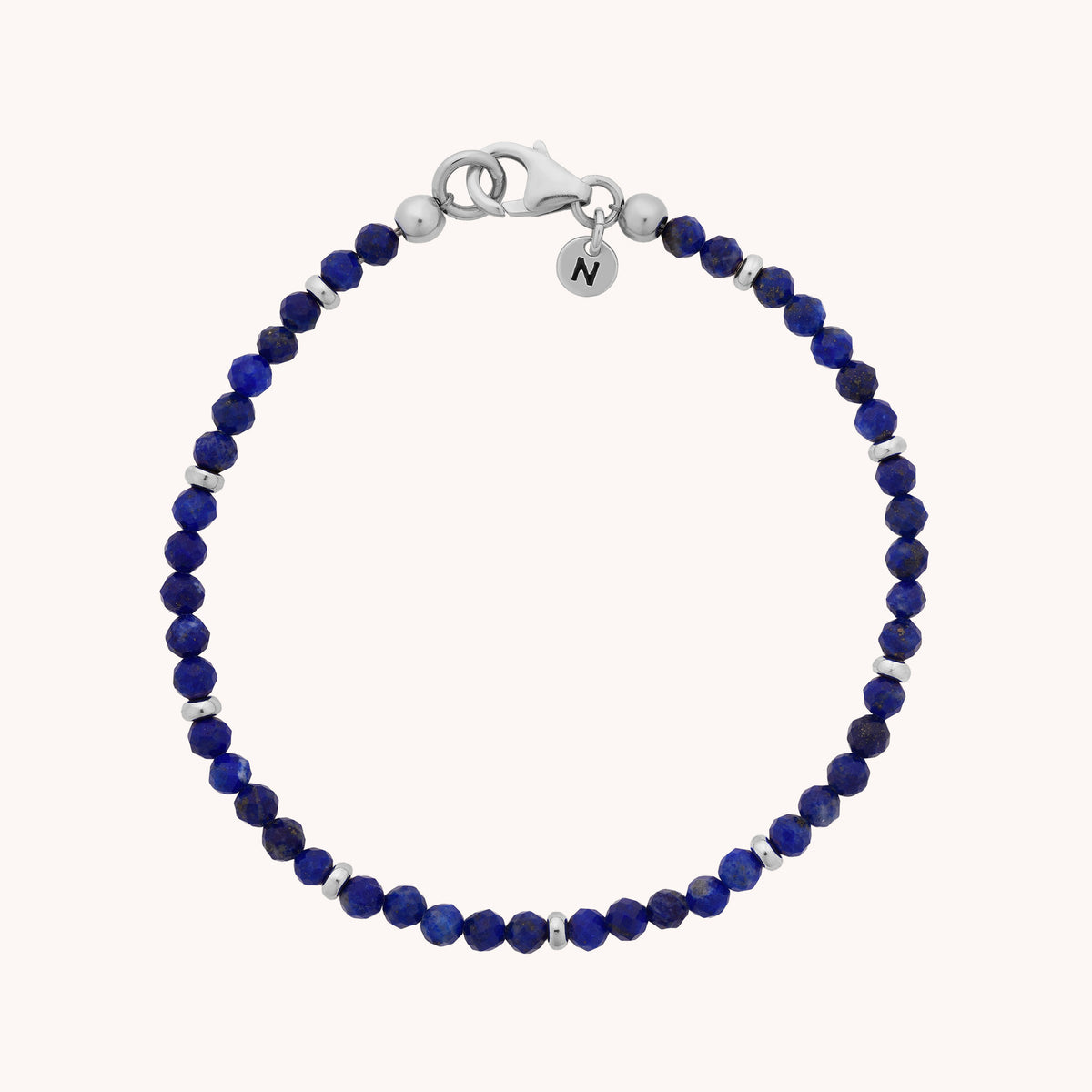 The Lapis Orbit Bracelet - September features deep blue and silver beads of genuine lapis lazuli, accented with a silver clasp and a delicate round charm engraved with the letter N. Presented on a plain background, this gemstone bracelet elegantly highlights the rich hues of lapis lazuli, making it a refined accessory and an ideal September birthstone gift.