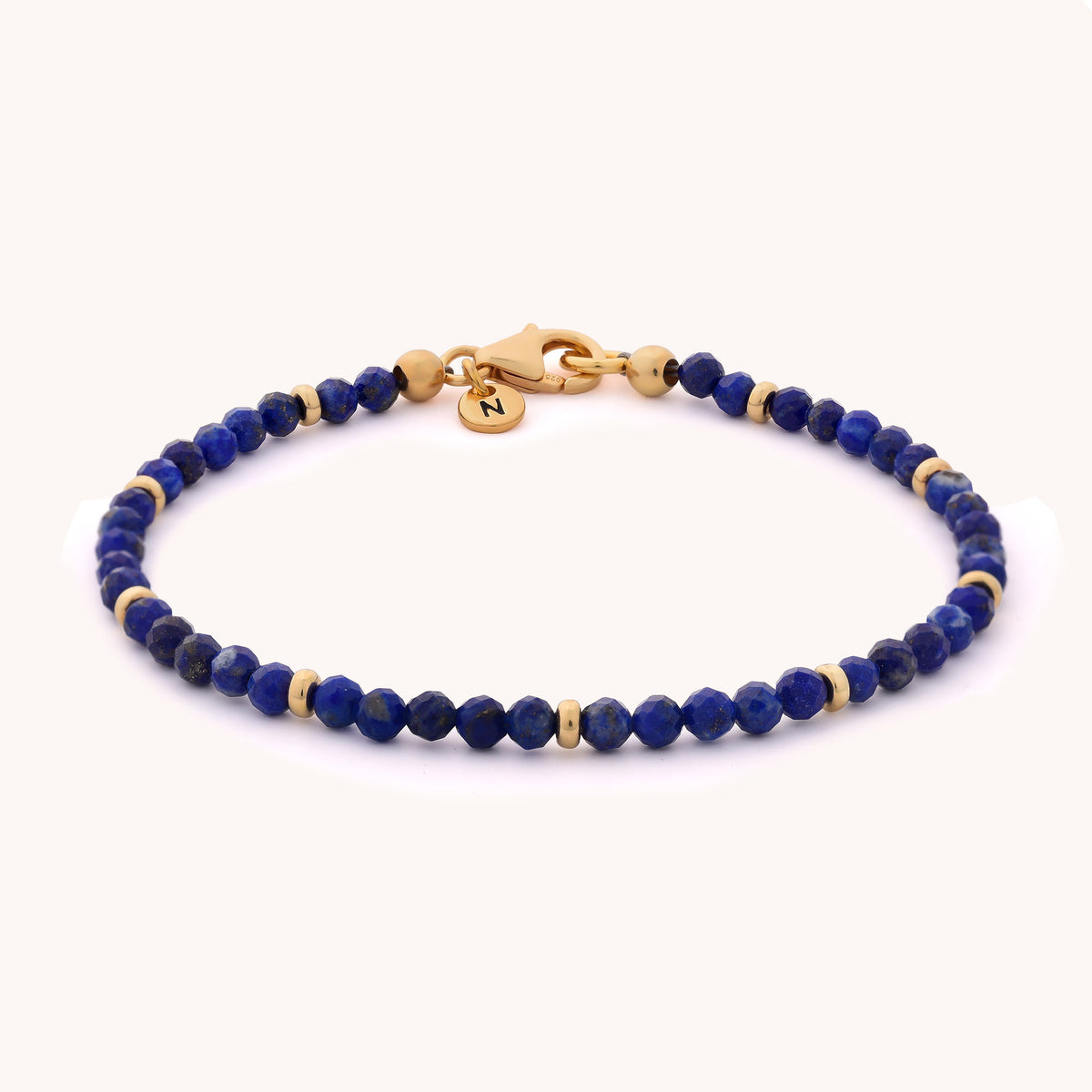 Introducing the Lapis Orbit Bracelet - September: A delicate gemstone bracelet adorned with small round lapis lazuli beads in a vibrant blue hue, accented by gold details. It features a gold clasp and a petite gold charm engraved with the letter Z, making it a perfect birthstone piece for September.