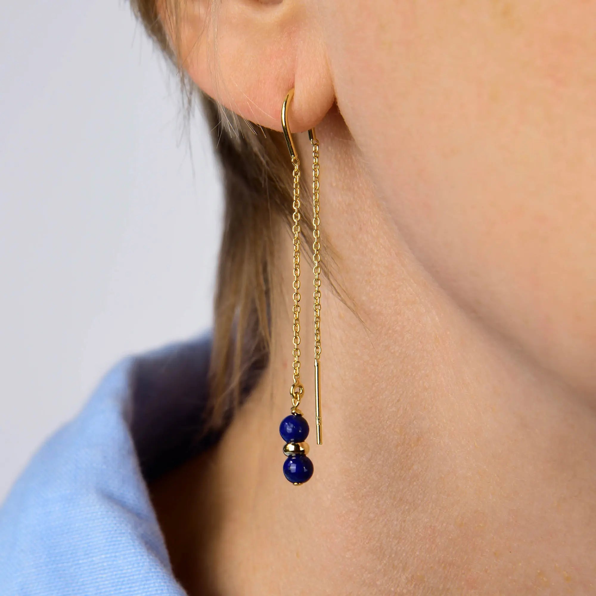 Close-up of a person wearing the Lapis Fall Earrings Birthstone September—elegant gold threader earrings featuring two small lapis-blue beads—shown against light skin and a blue collared shirt.