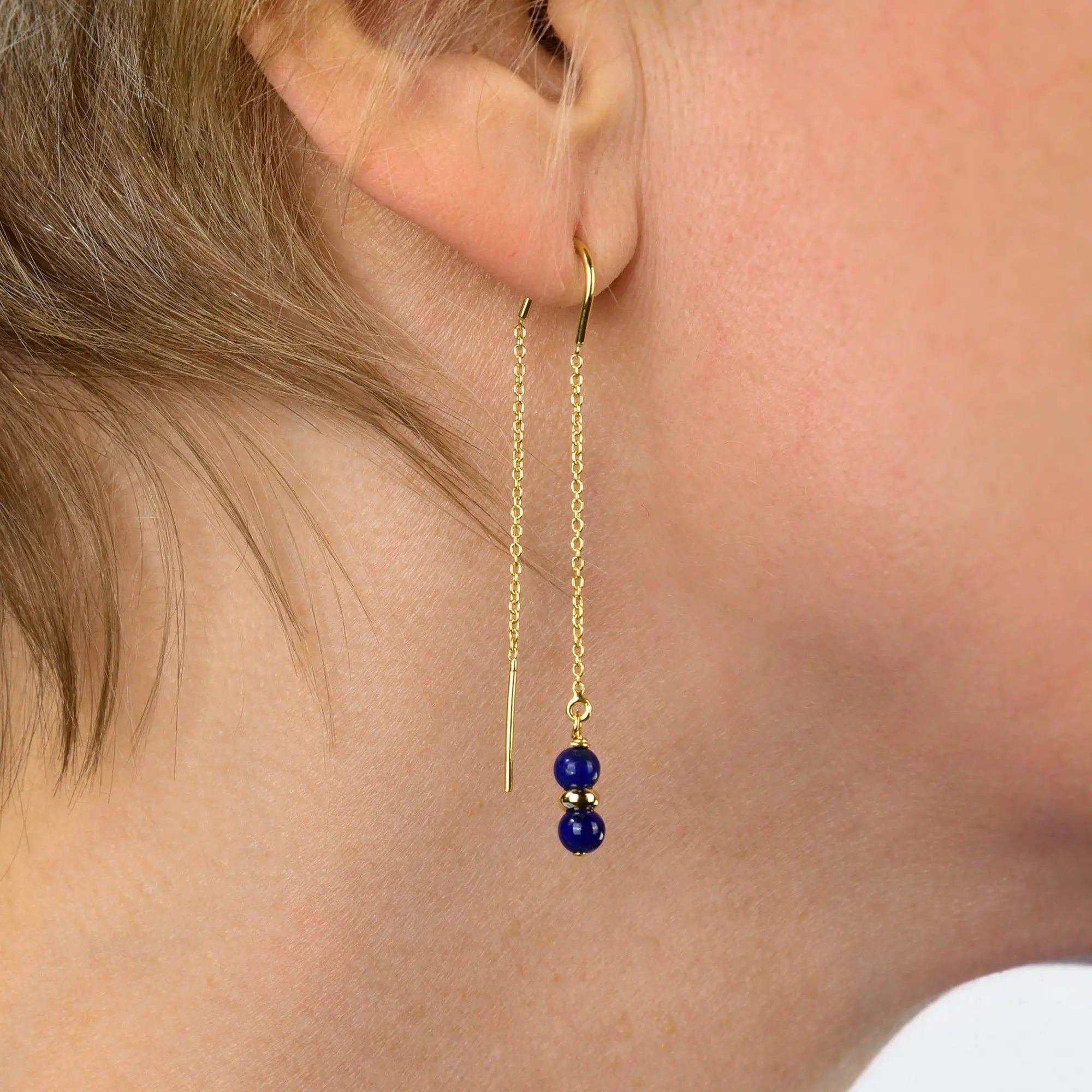 Close-up of a person’s ear wearing the Lapis Fall Earrings Birthstone September—a gold chain threader earring featuring two small lapis-inspired blue beads separated by a gold spacer, set against a light background.