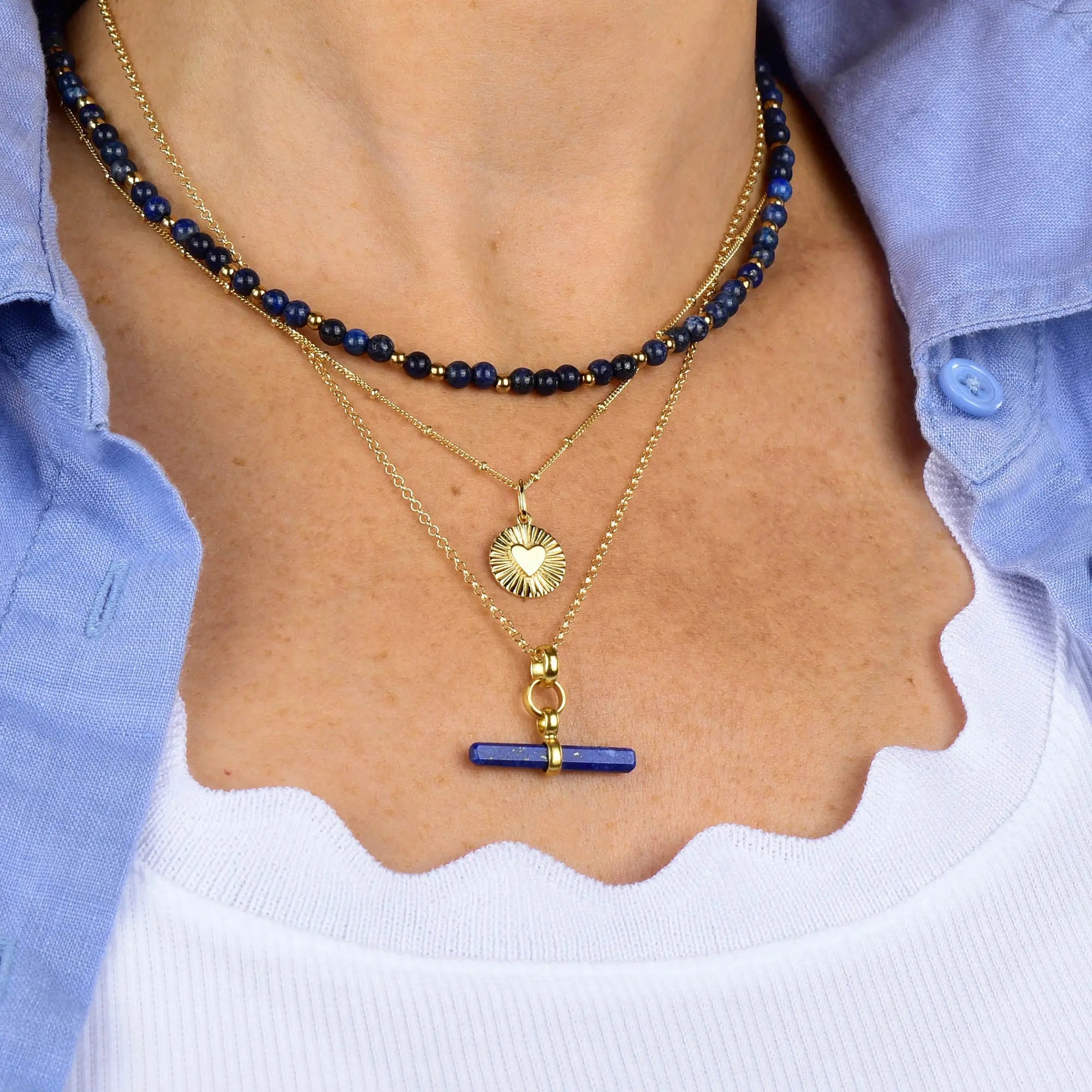A person wearing layered gold plated necklaces with blue beads and pendants, including the Shining Heart Necklace Sterling Silver, over a white top and light blue shirt.
