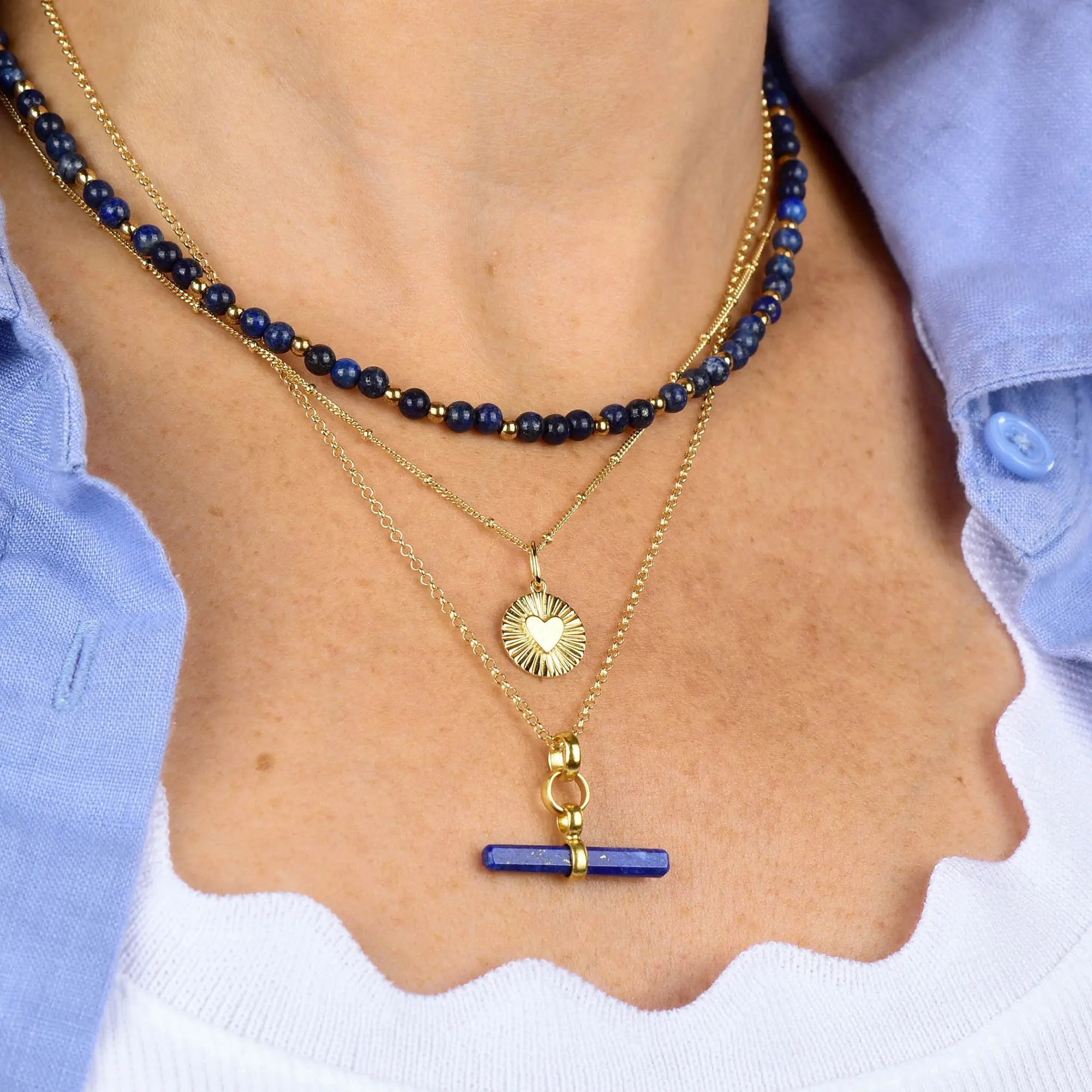 A person wearing three layered necklaces, including the Shining Heart Necklace Sterling Silver, all styled over a white top and blue shirt.