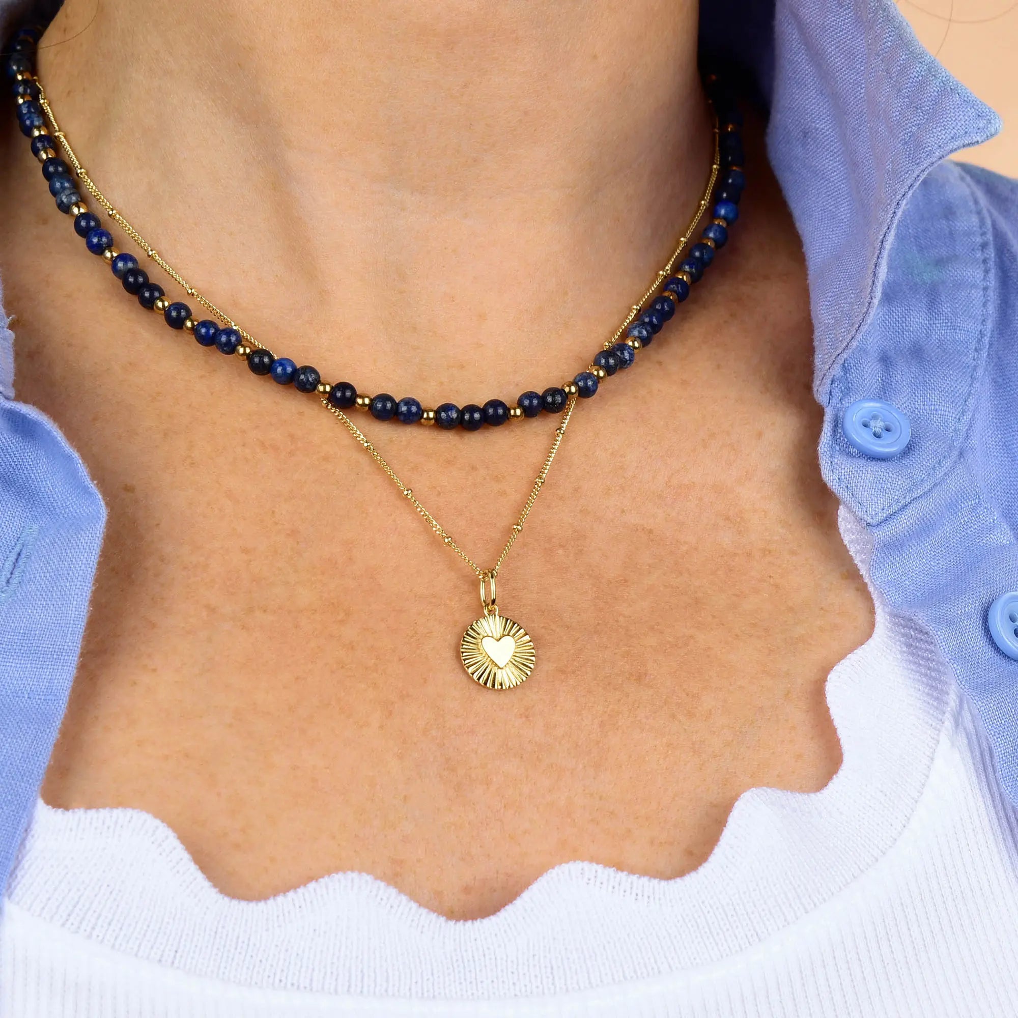 A close-up of a person wearing two necklaces: a blue beaded choker and the Shining Heart Necklace Sterling Silver, featuring a sunburst heart pendant. The person is dressed in a white top and a blue button-up shirt.