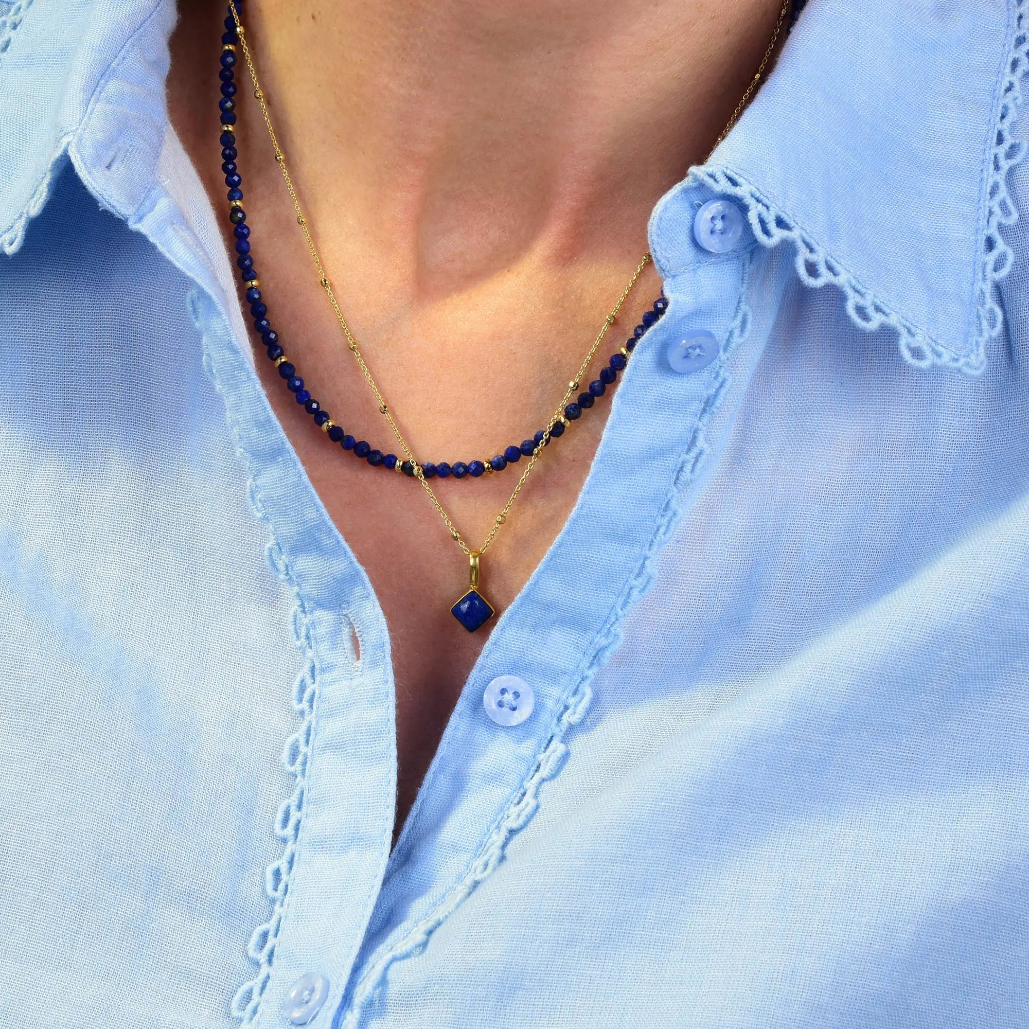 A person wearing a light blue shirt with lacy edges and two necklaces: one is an eco silver chain adorned with small blue beads, while the other is a gold Lapis Diamond Birthstone Necklace for September featuring a striking blue gemstone pendant.