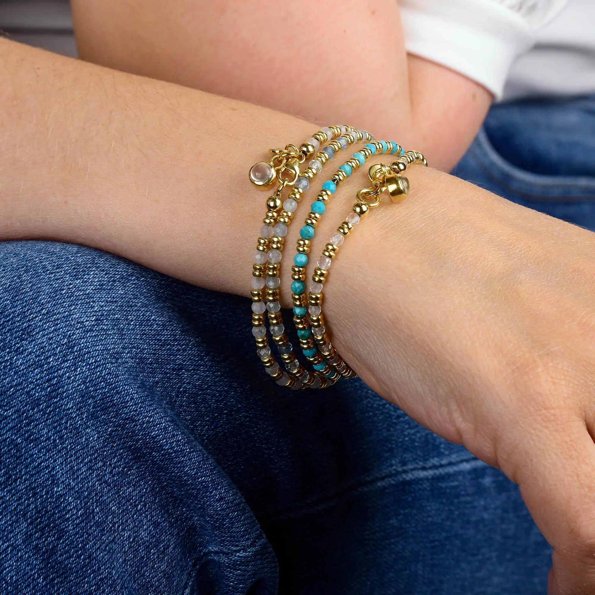 A close-up of a person’s wrist adorned with layered beaded bracelets in gold, turquoise, and white tones, including the stunning Clear Quartz Linear Birthstone Bracelet for April. The person is dressed in blue jeans and a white shirt.