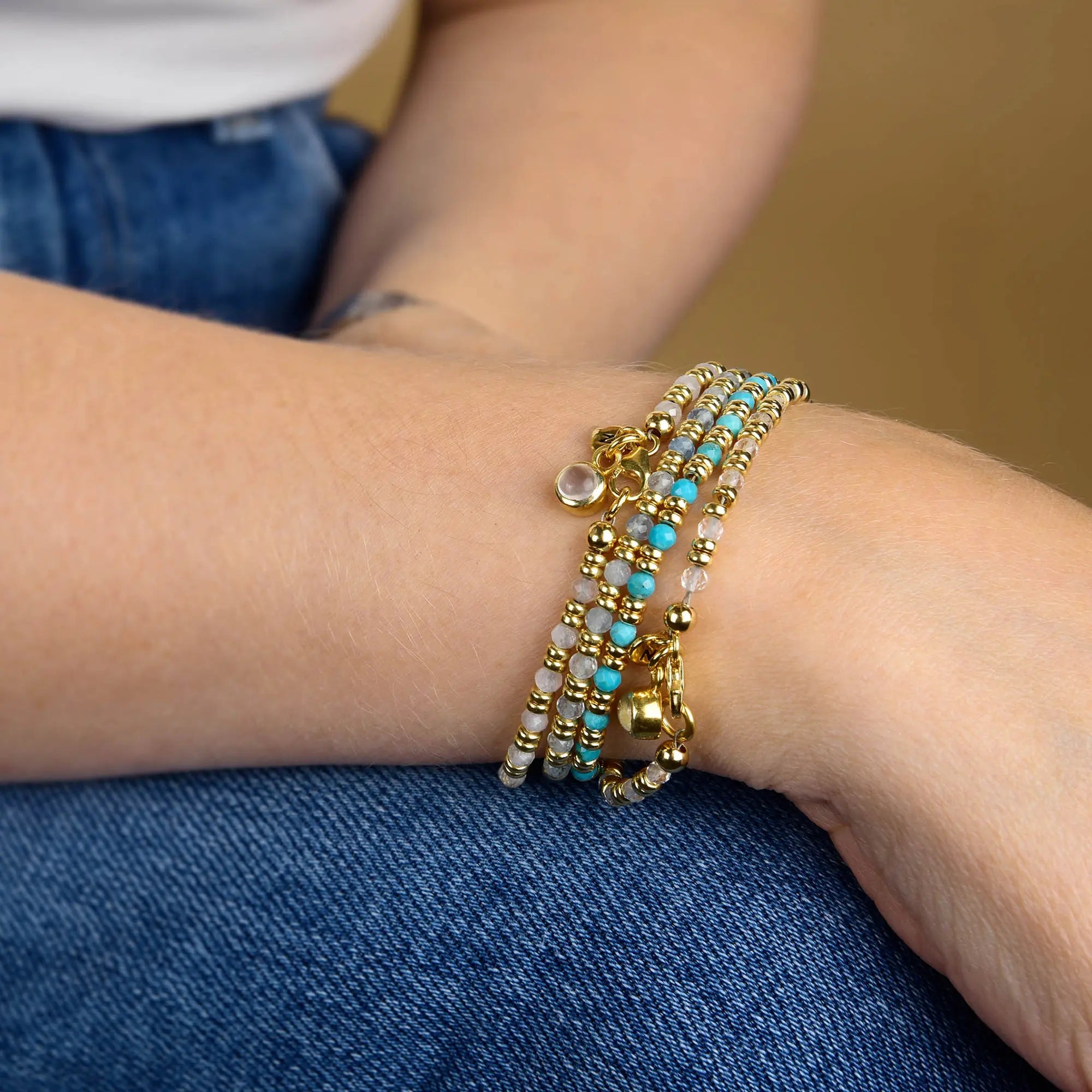 A person wearing blue jeans and a white shirt has their arms crossed, showcasing multiple Linear Birthstone Bracelets with gold, turquoise, and white gemstone beads on their wrist.