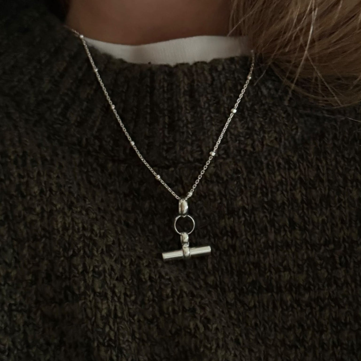 A person wearing a dark, textured sweater and the Mini T Bar Necklace featuring a circle and bar toggle pendant on a gold-plated sterling silver satellite chain.