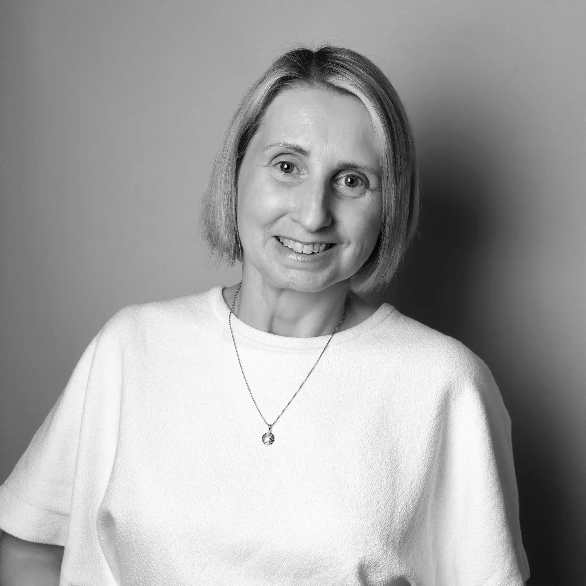 Smiling woman with short, straight blonde hair wearing a white textured short-sleeve top and the Saint Christopher Necklace on a sterling silver chain, standing against a plain background in a black and white photo.