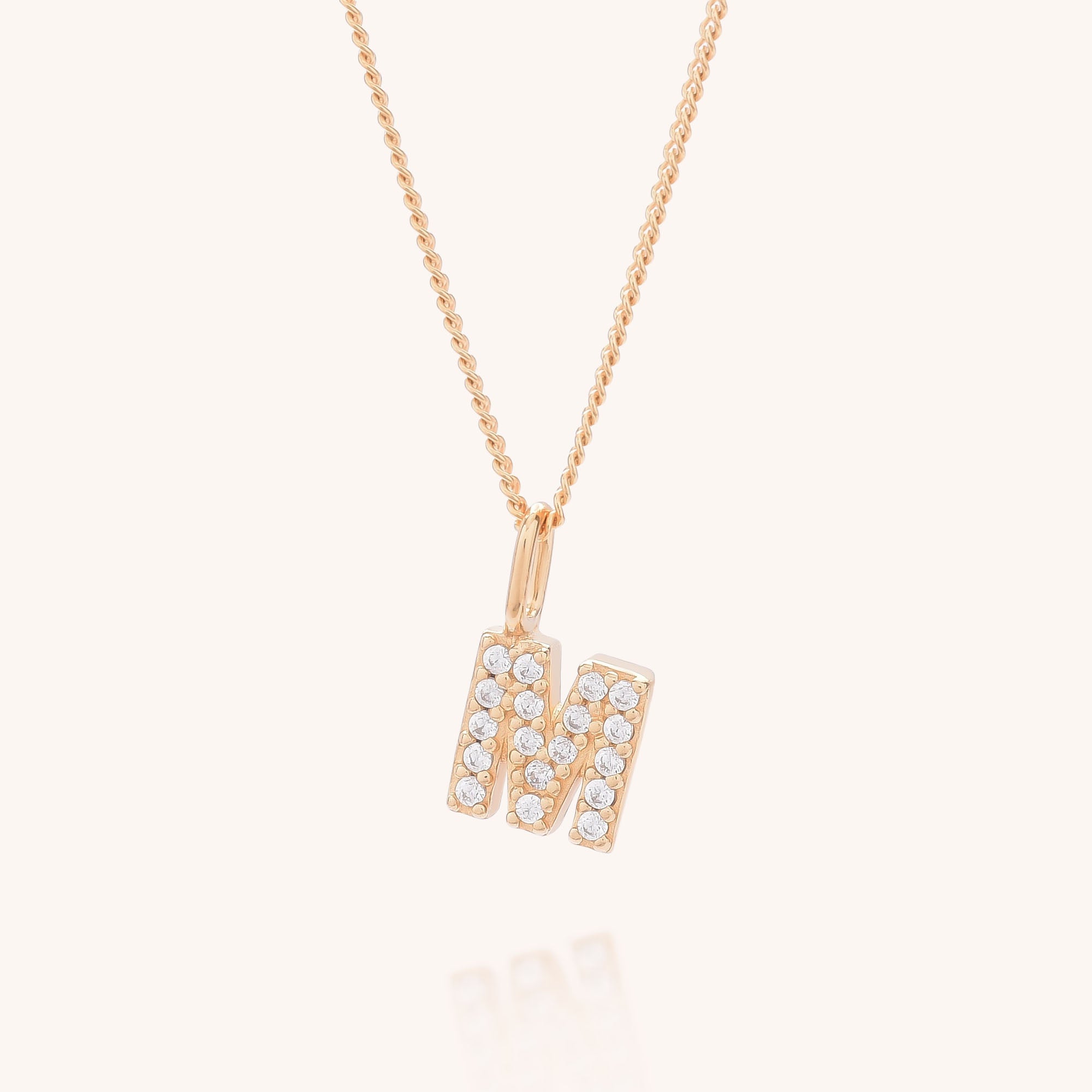 The Initial Sparkle Necklace features a gold pendant shaped like the letter M, accented with shimmering cubic zirconia, and is shown against a light background.
