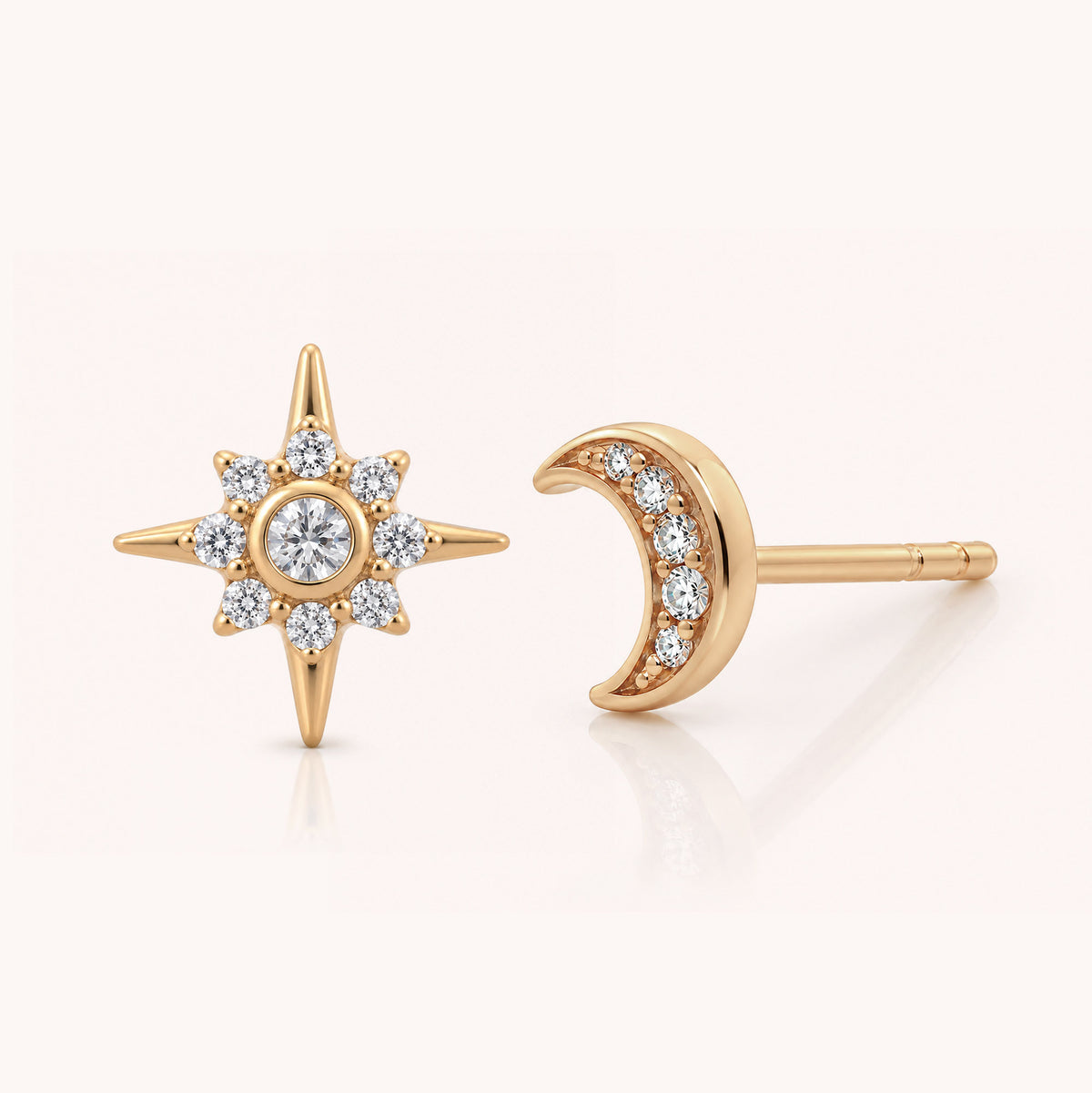 A pair of gold celestial stud earrings, featuring a starburst design with a central round diamond surrounded by six smaller diamonds, and a crescent moon adorned with three small diamonds, shown on a light cream background.