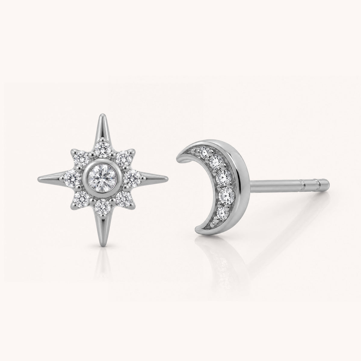 A pair of silver-toned stud earrings, one shaped like a six-pointed star with a central round clear gemstone surrounded by smaller stones, and the other as a crescent moon with a row of clear stones along the curve, shown on a light cream background.