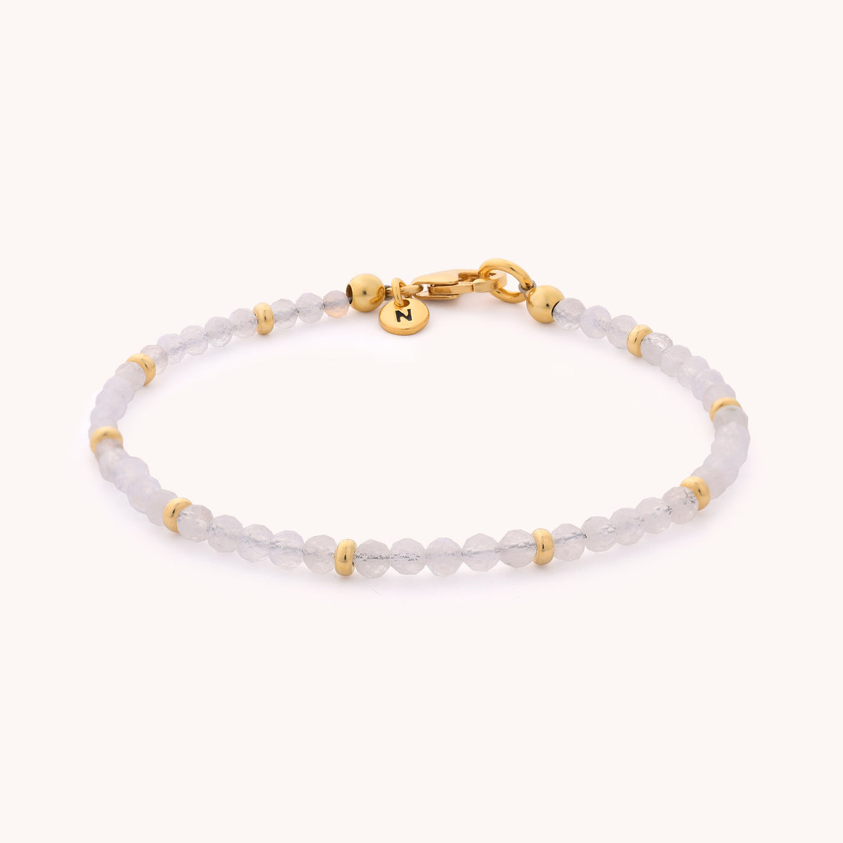 Introducing the Rainbow Moonstone Orbit Bracelet - June: a delicate, handcrafted bracelet featuring translucent white beads reminiscent of rainbow moonstone, accented with small gold spacer beads. The design is completed with a gold clasp and a round gold charm engraved with the letter N.
