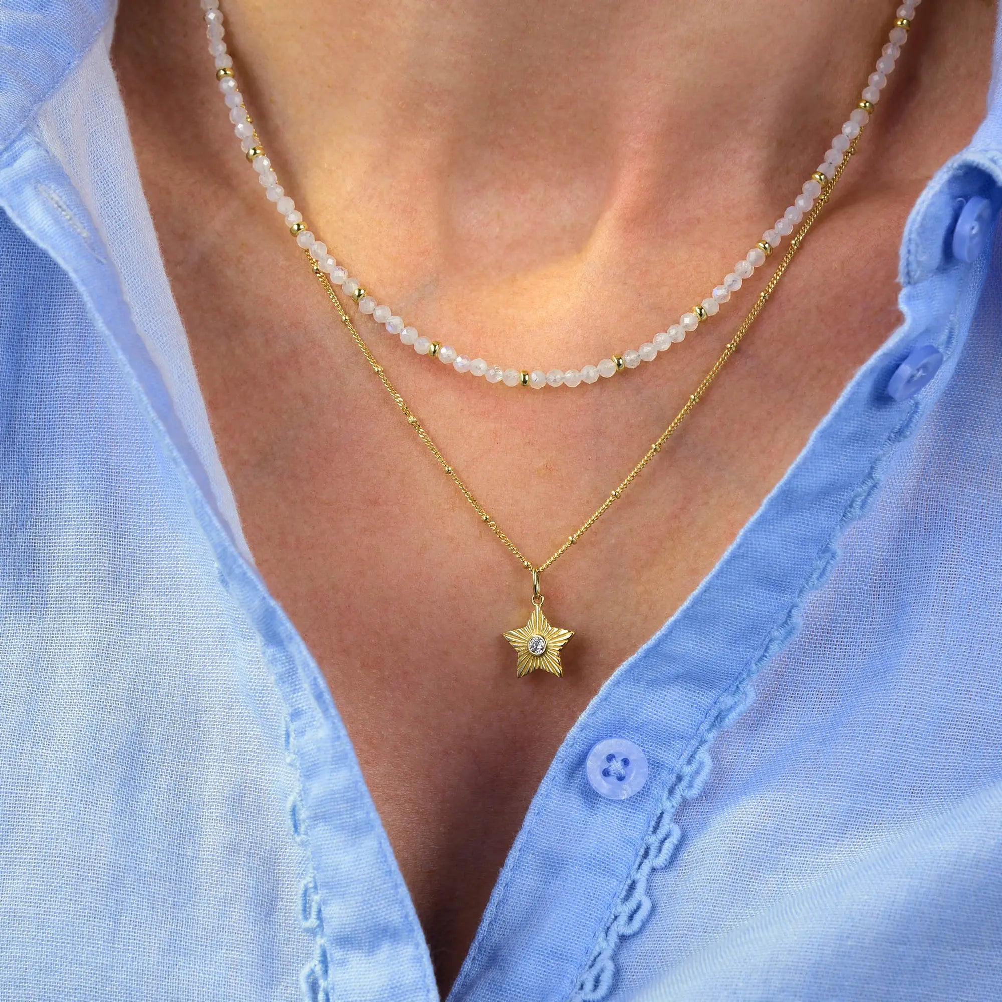 A person wearing a light blue shirt with two necklaces: a beaded choker and the Astrid Star Necklace—a gold, sterling silver piece featuring a sparkling cubic zirconia gemstone in the center.