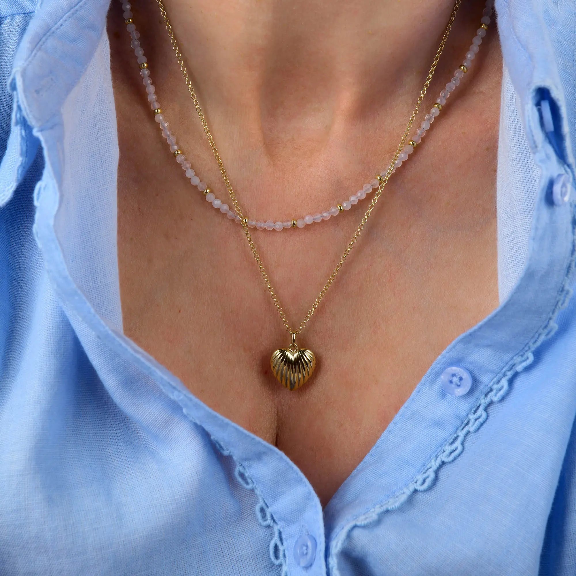A person wearing a light blue shirt with two layered necklaces: one is the Scallop Heart Necklace featuring a gold heart pendant, and the other is adorned with small pink beads.