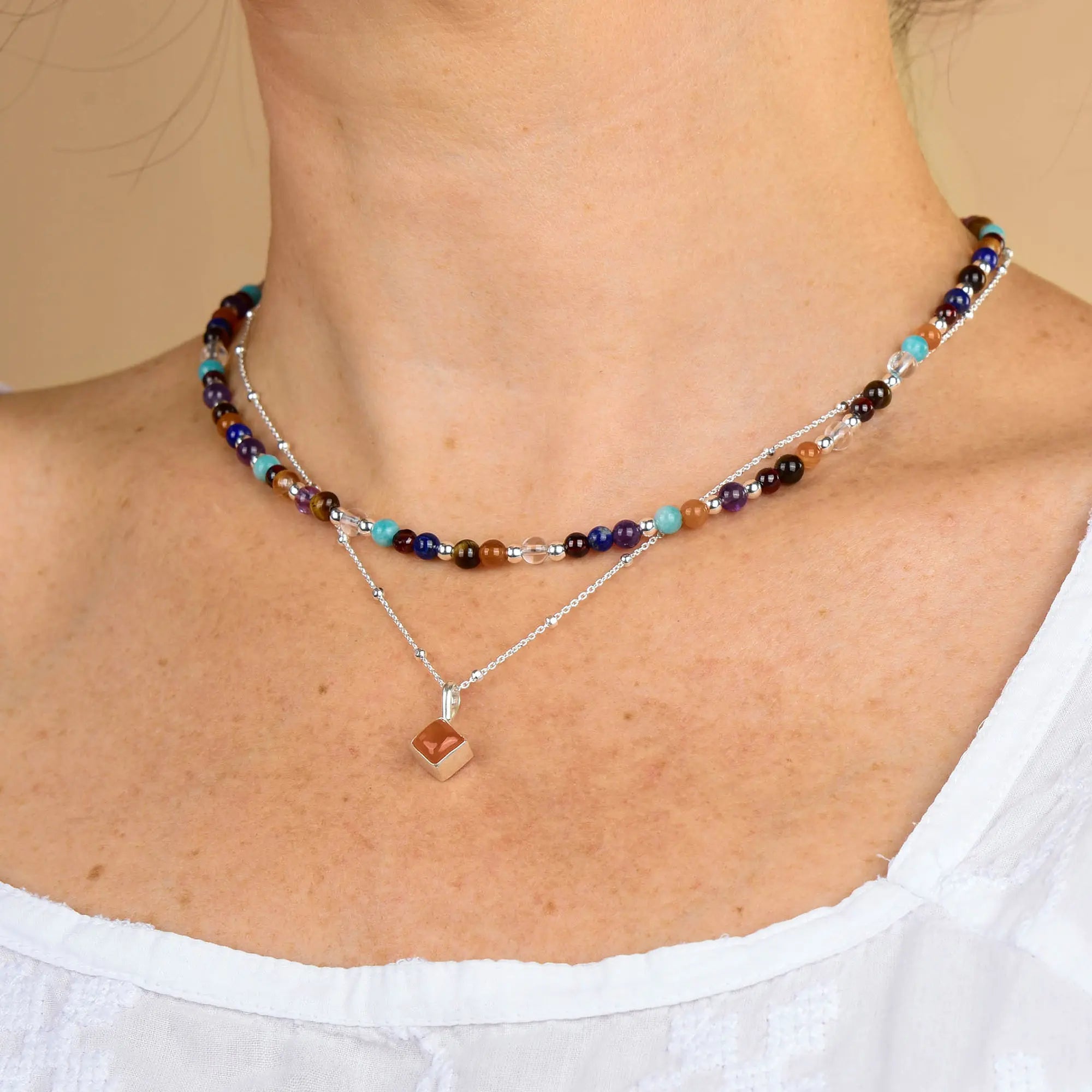 A person wearing the Multi Gemstone Necklace, a double-layered beaded piece with colorful gemstones, paired with a sterling silver chain necklace featuring a small cube pendant and a white, textured top.