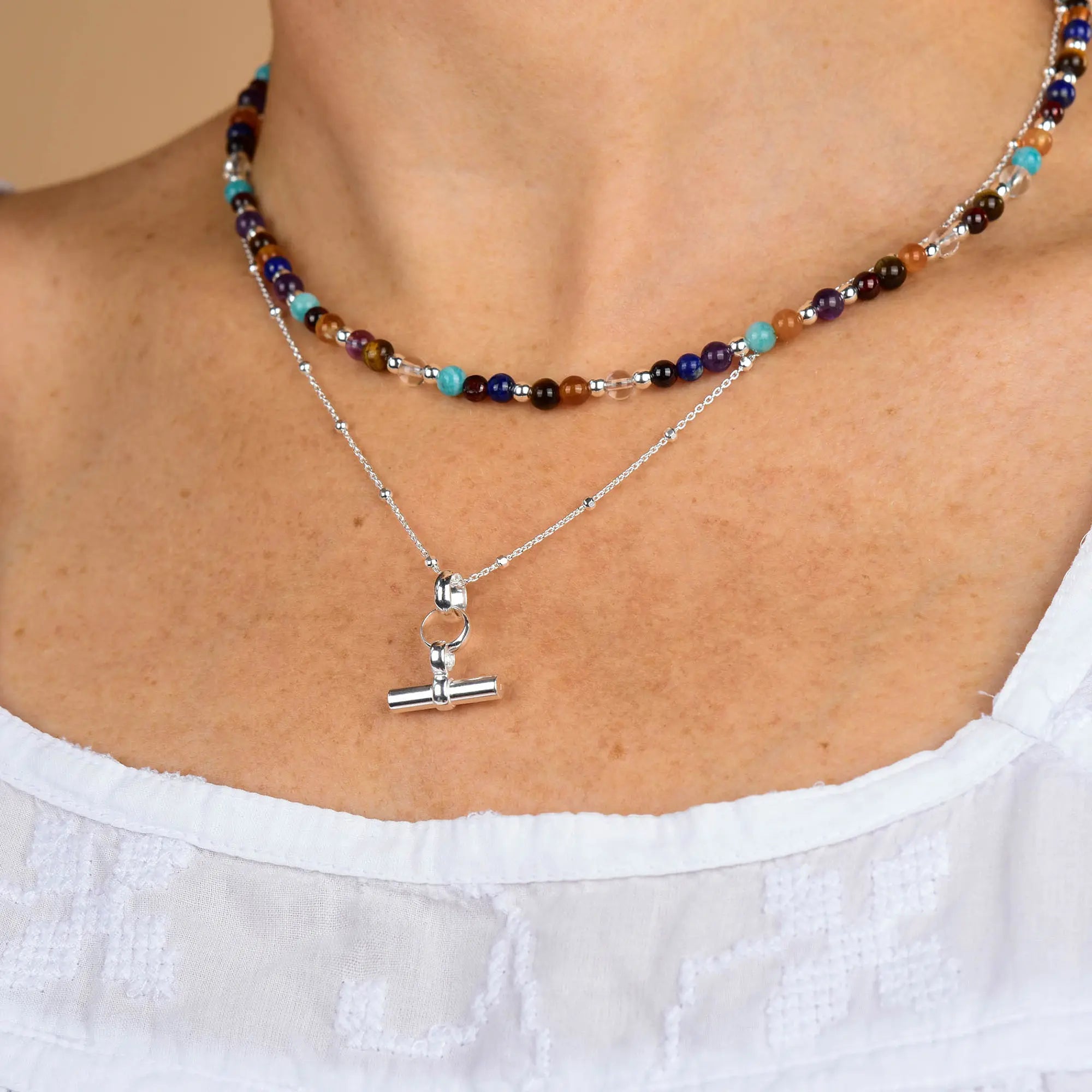 A close-up of a person wearing two necklaces: a colorful beaded necklace and the Mini T Bar Necklace—a sterling silver satellite chain with a T-bar pendant—over a white, textured, off-shoulder top.