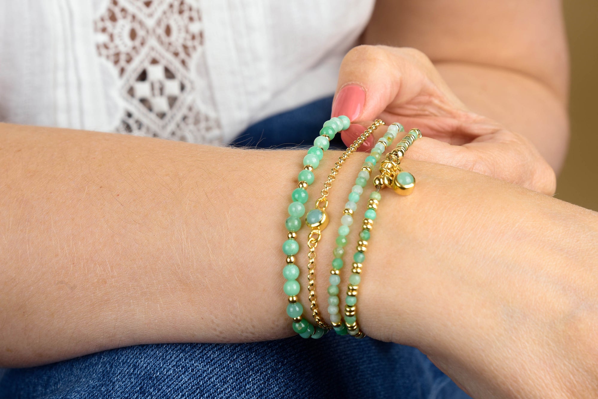 A person wearing a white top and blue jeans fastens the Chrysoprase Jewel Bead Birthstone Bracelet – May, featuring green beads and gold accents, around their wrist. With pink-painted nails, this accessory makes an ideal May birthstone gift.