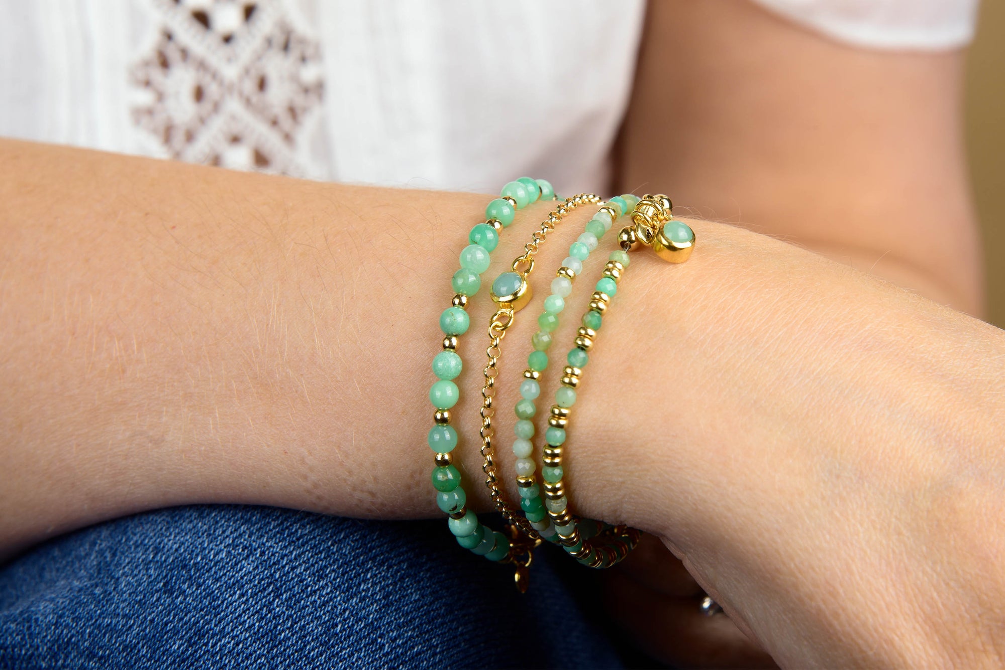 A person wearing multiple turquoise and gold beaded bracelets, including the Chrysoprase Jewel Bead Birthstone Bracelet - May—a perfect gift for those with a May birthday—paired with a white lace top and blue jeans.
