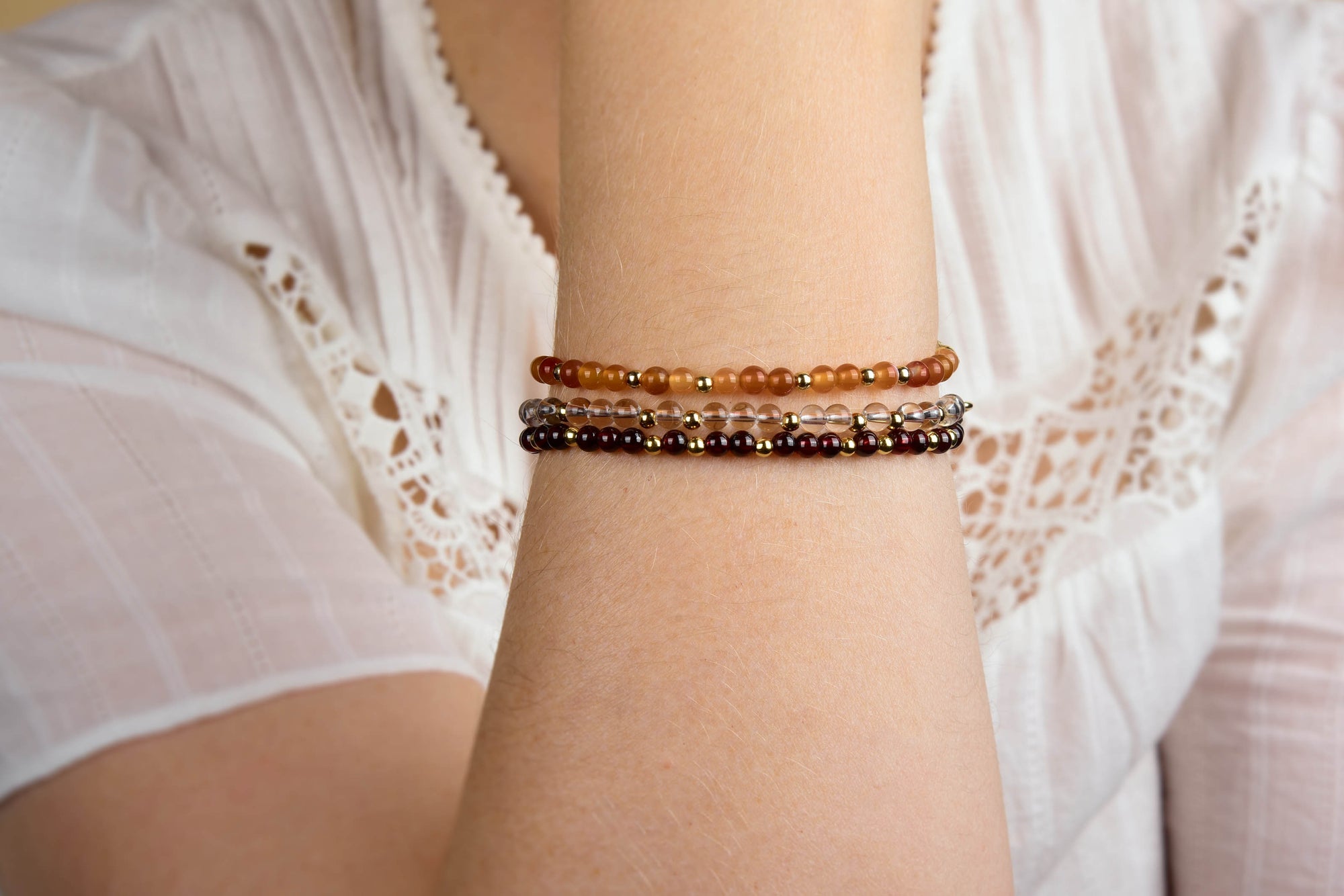 A person wearing a white, short-sleeve, lace-trimmed blouse rests their chin on their hand, displaying two earth-toned beaded bracelets and the Garnet Jewel Bead Birthstone Bracelet – January, featuring delicate garnet beads with sterling silver accents, on their wrist.