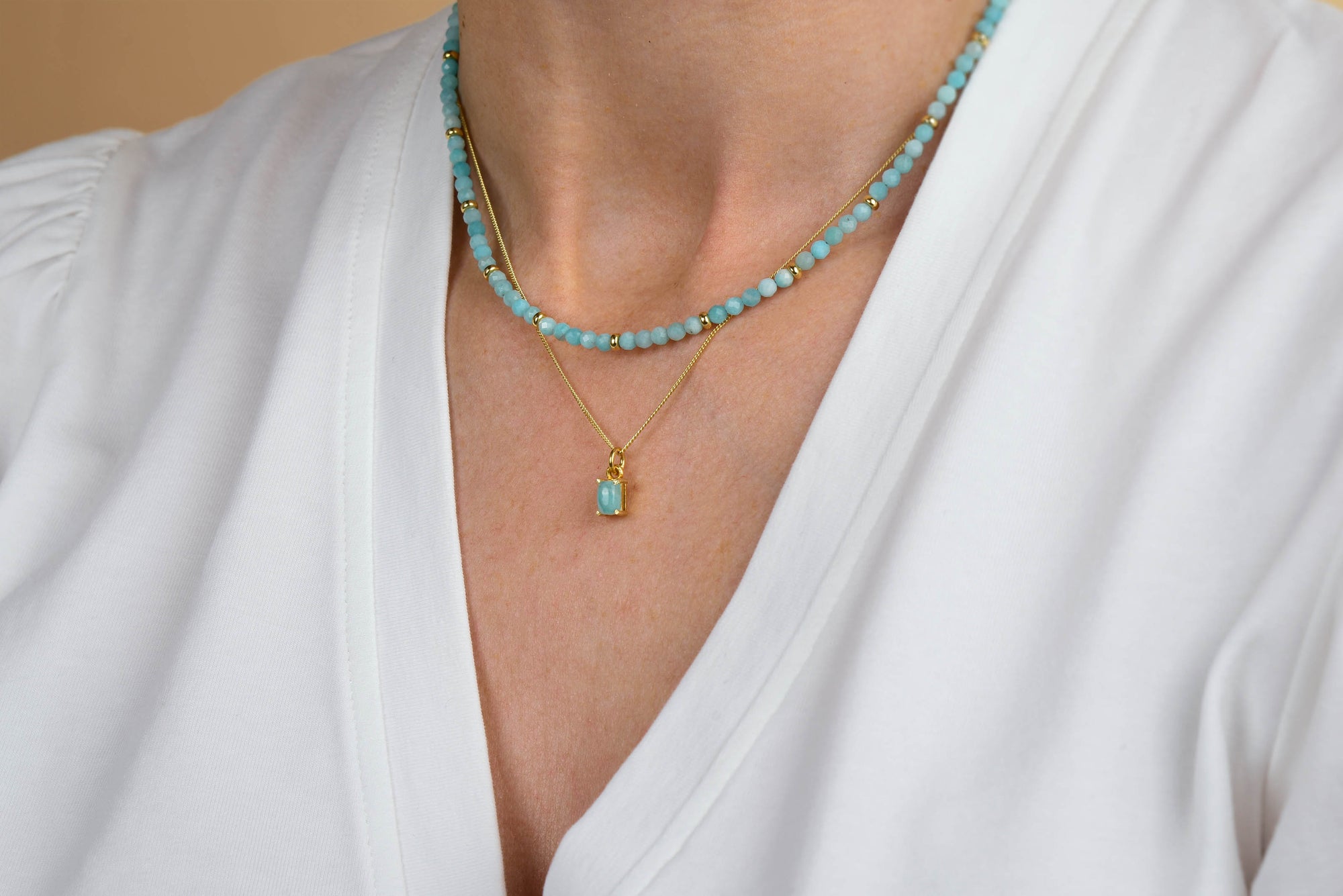A person wearing a white V-neck shirt, with two turquoise bead necklaces layered on their neck—one featuring the Amazonite Octavia Necklace, distinguished by its octagonal design and gold pendant set with a turquoise stone.